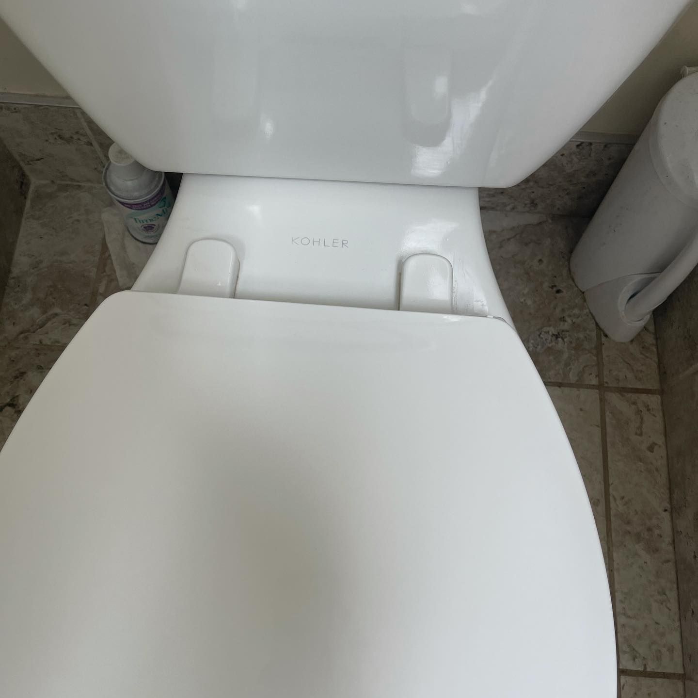 A top-down view of a closed white toilet seat and lid against a tiled floor, with a cleaning bottle near the tank.