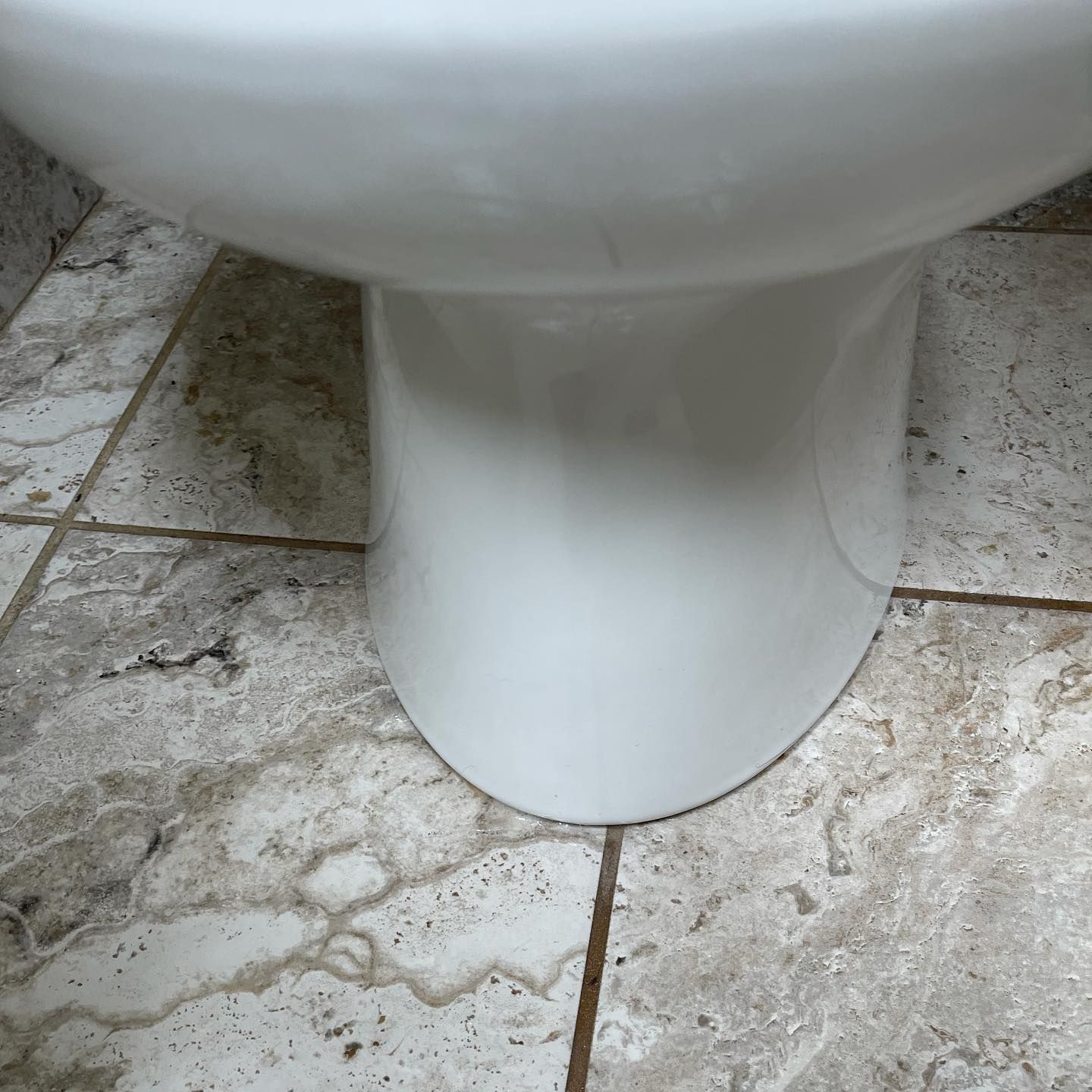 A close-up view of the white porcelain base of a toilet resting on a light-colored, mottled tiled floor.