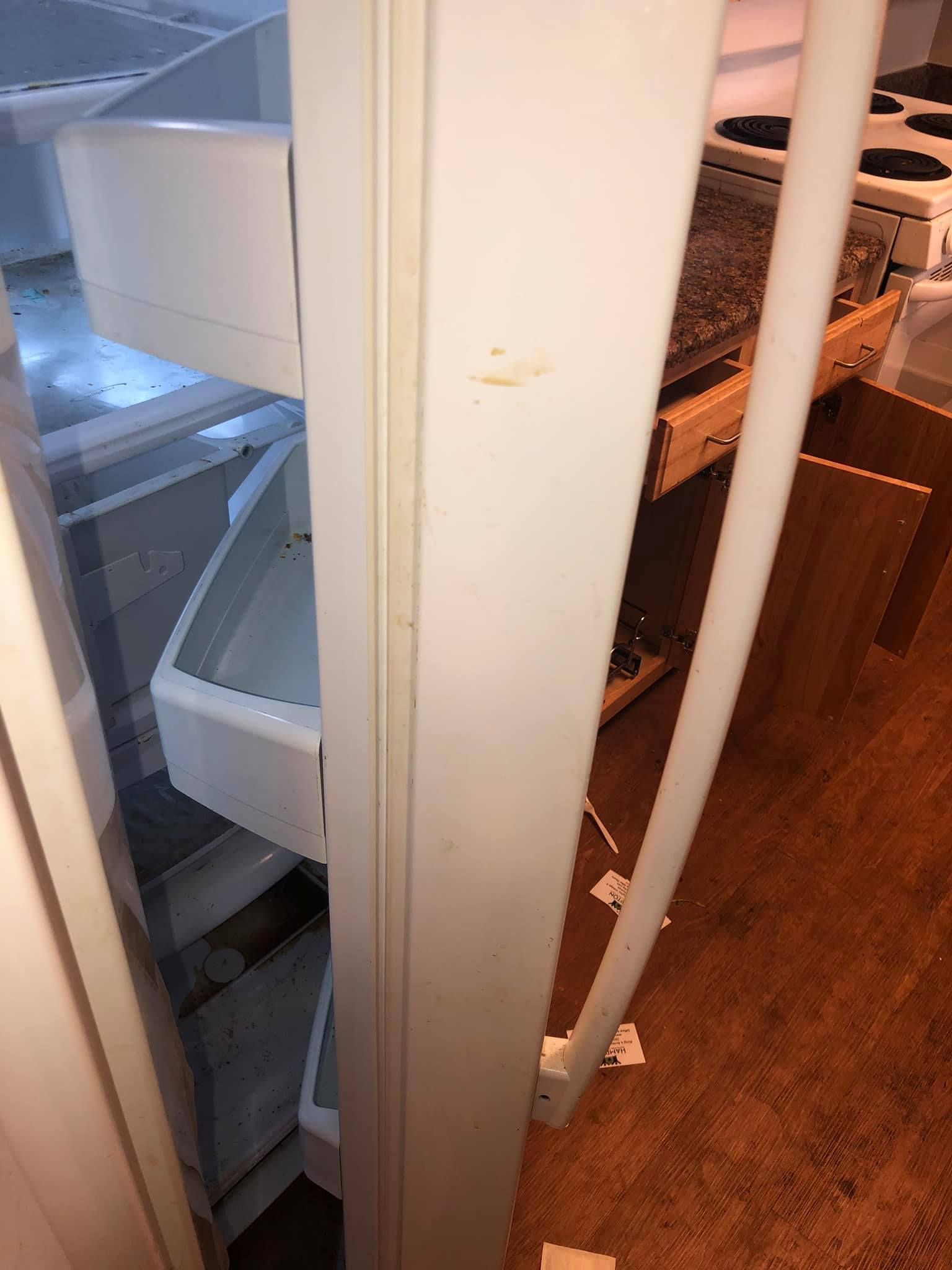 A white refrigerator door stands open, partially blocking the view of a kitchen stove and cabinets on a wooden floor.
