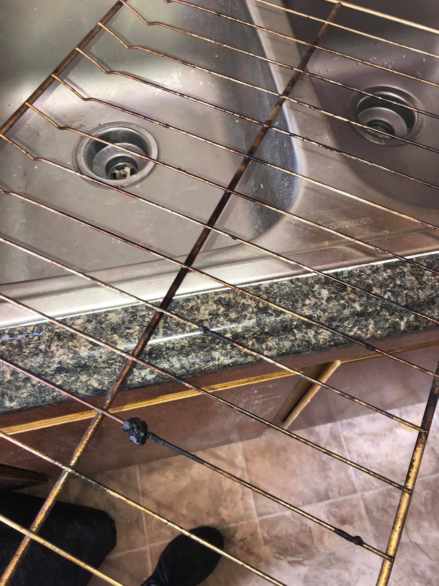 A metal oven rack resting over a double kitchen sink with dark, grimy buildup on its wires.