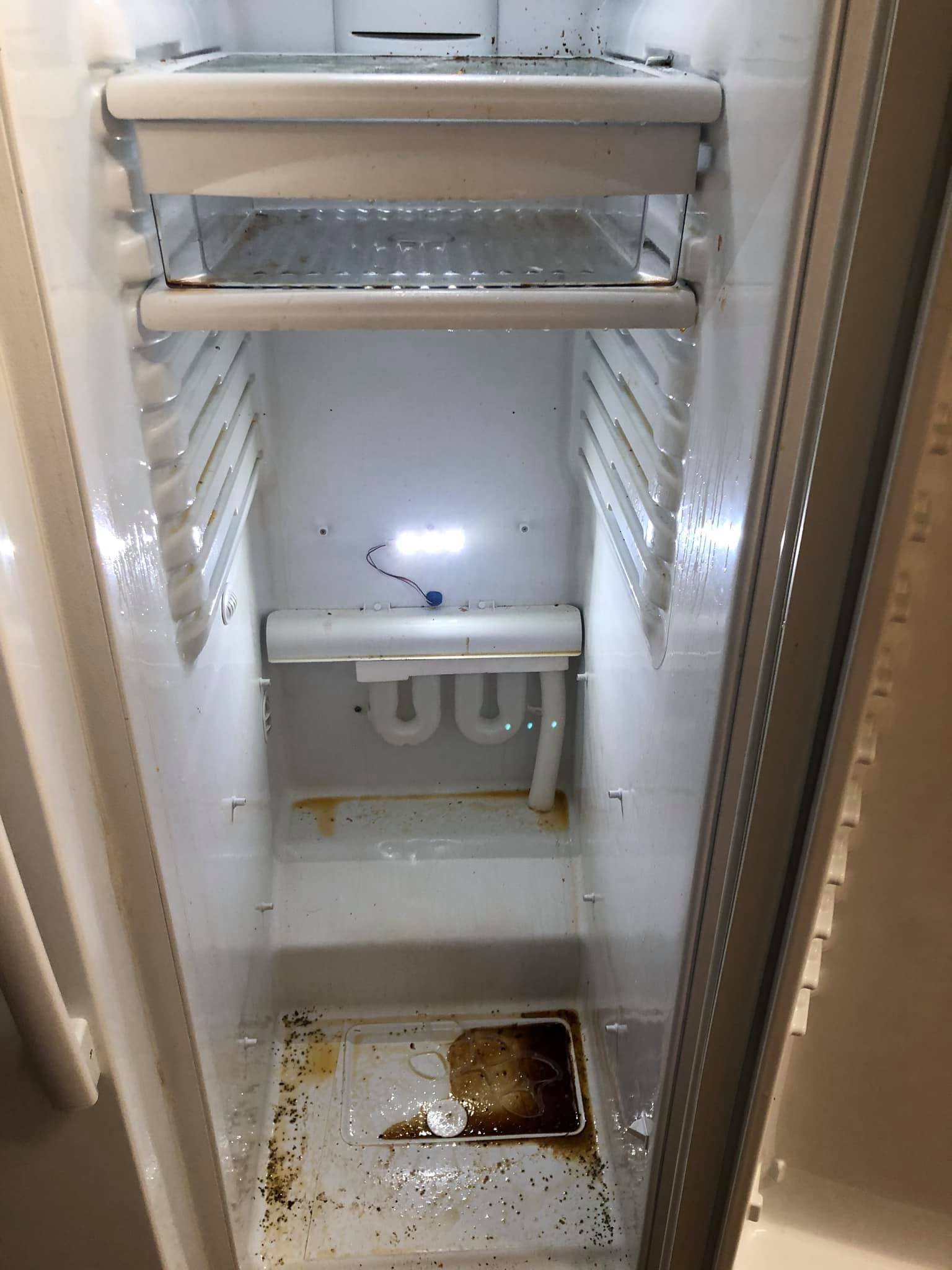 The interior of an empty, dirty white refrigerator with shelves, liquid residue, and debris on the floor.