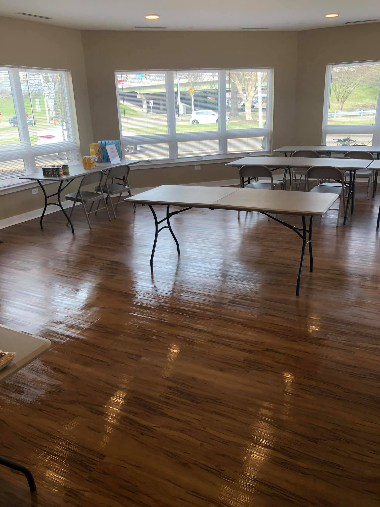 A bright room with wood flooring, large windows, and multiple folding tables and chairs arranged for a meeting.