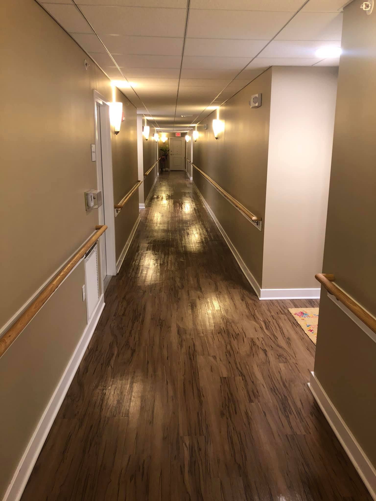 A long, dimly lit indoor hallway with wooden floors, tan walls, chair rails, and handrails on both sides.