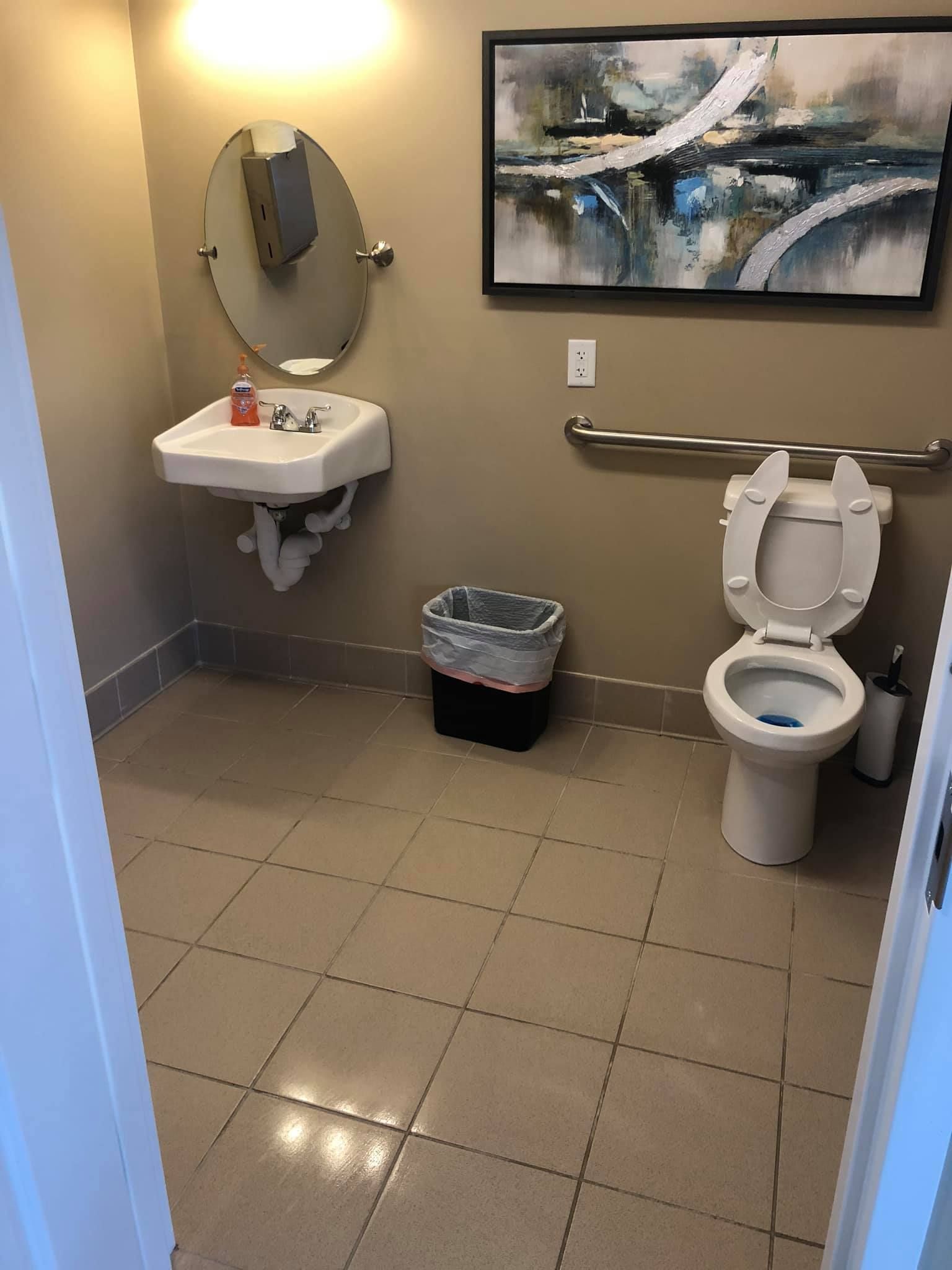 A modern, neutral-toned restroom featuring a wall-mounted sink, a toilet with a grab bar, and a framed abstract painting.