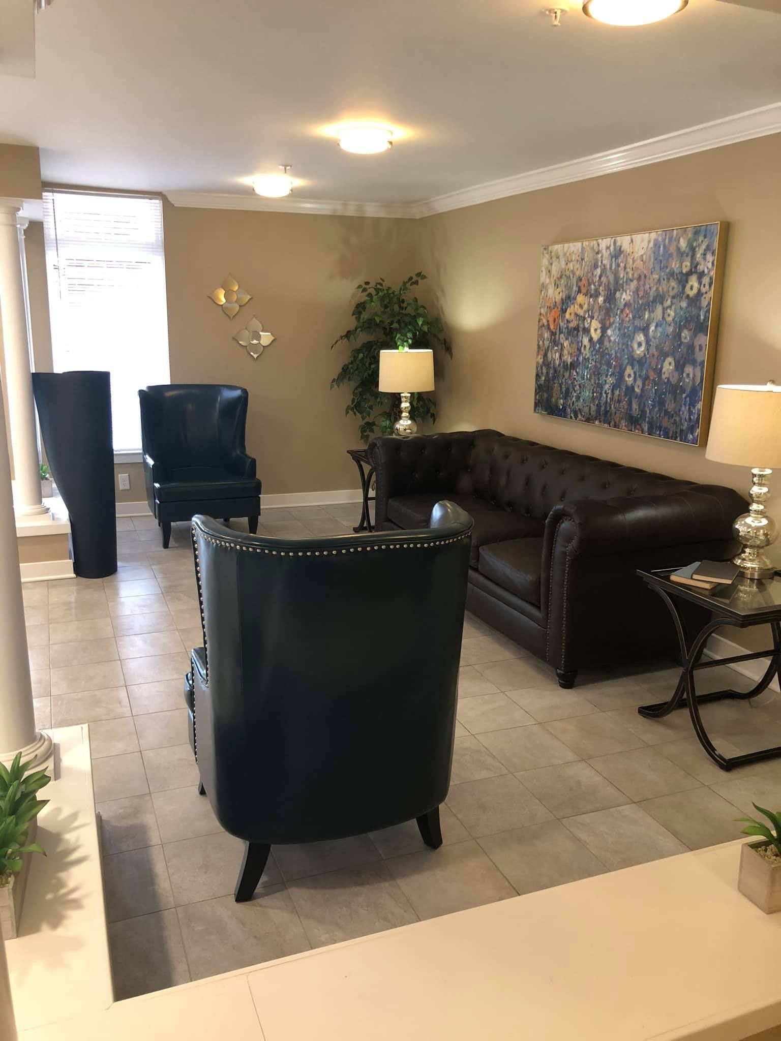 A furnished living area with a dark leather sofa, two matching armchairs, a side table, and a large abstract wall painting.