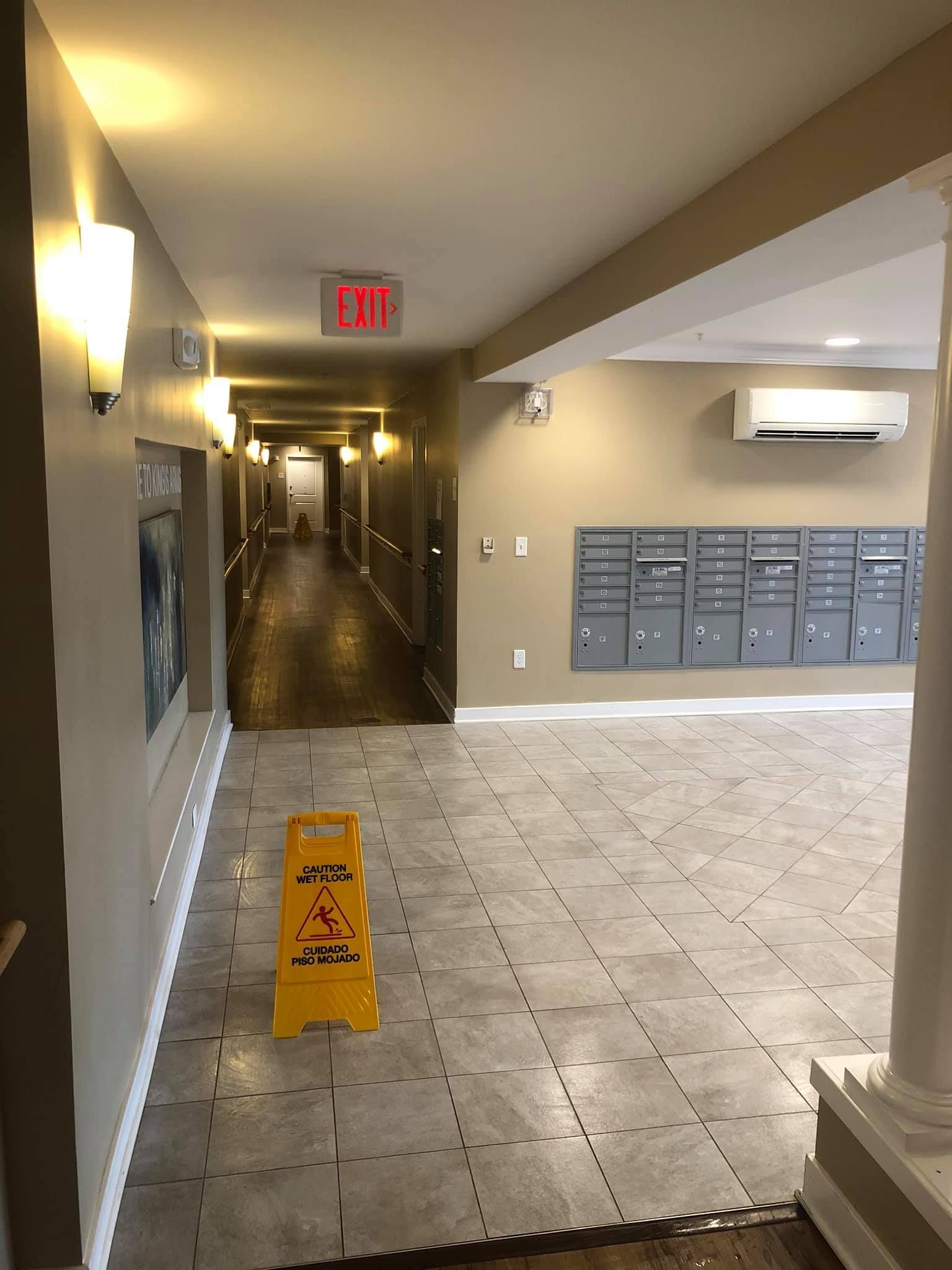 A hallway in an apartment building with a wet floor sign, mailboxes on the wall, and an exit sign overhead.
