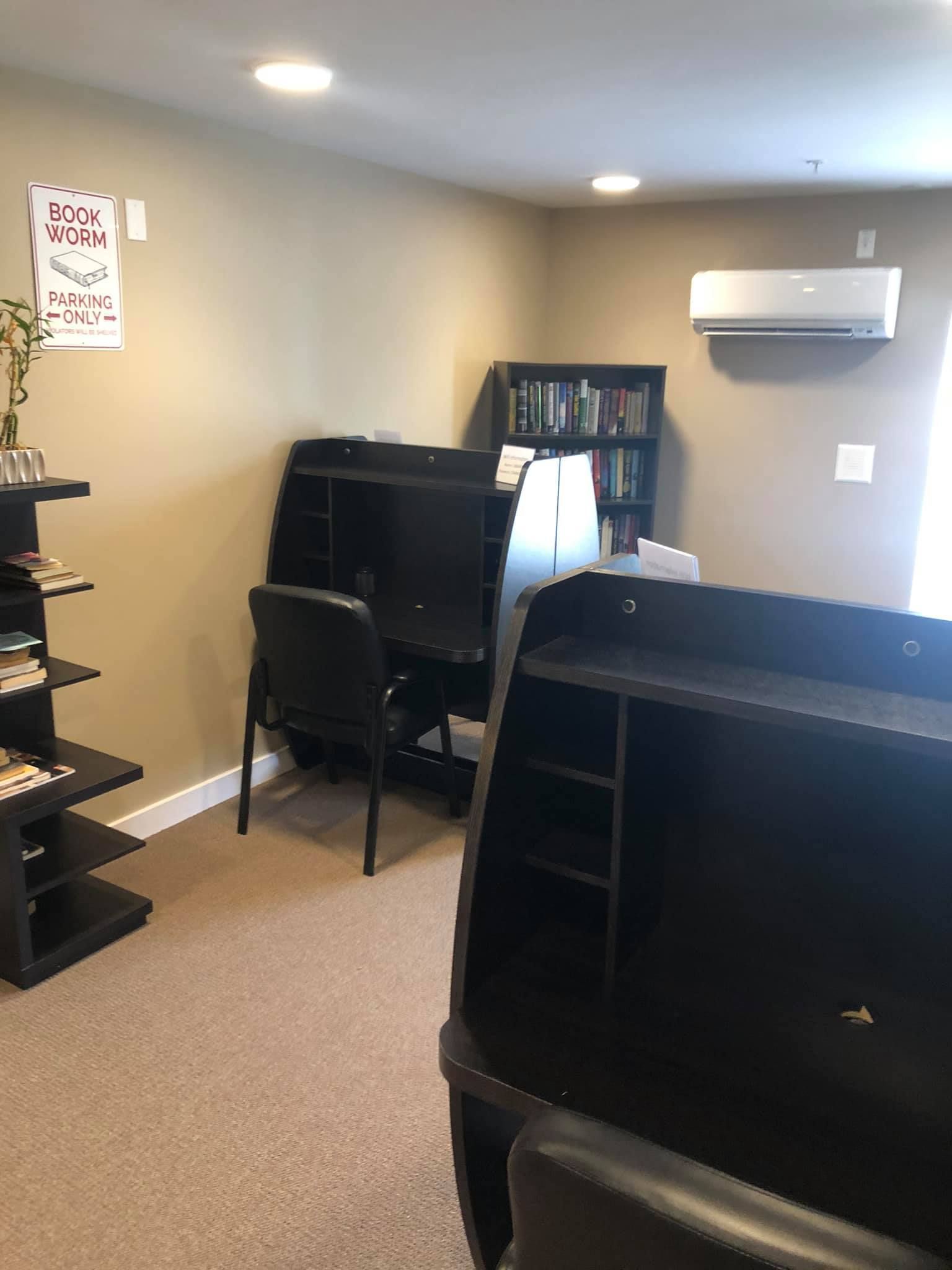 An office space with two black desks, matching chairs, a bookshelf, and a wall-mounted air conditioner.