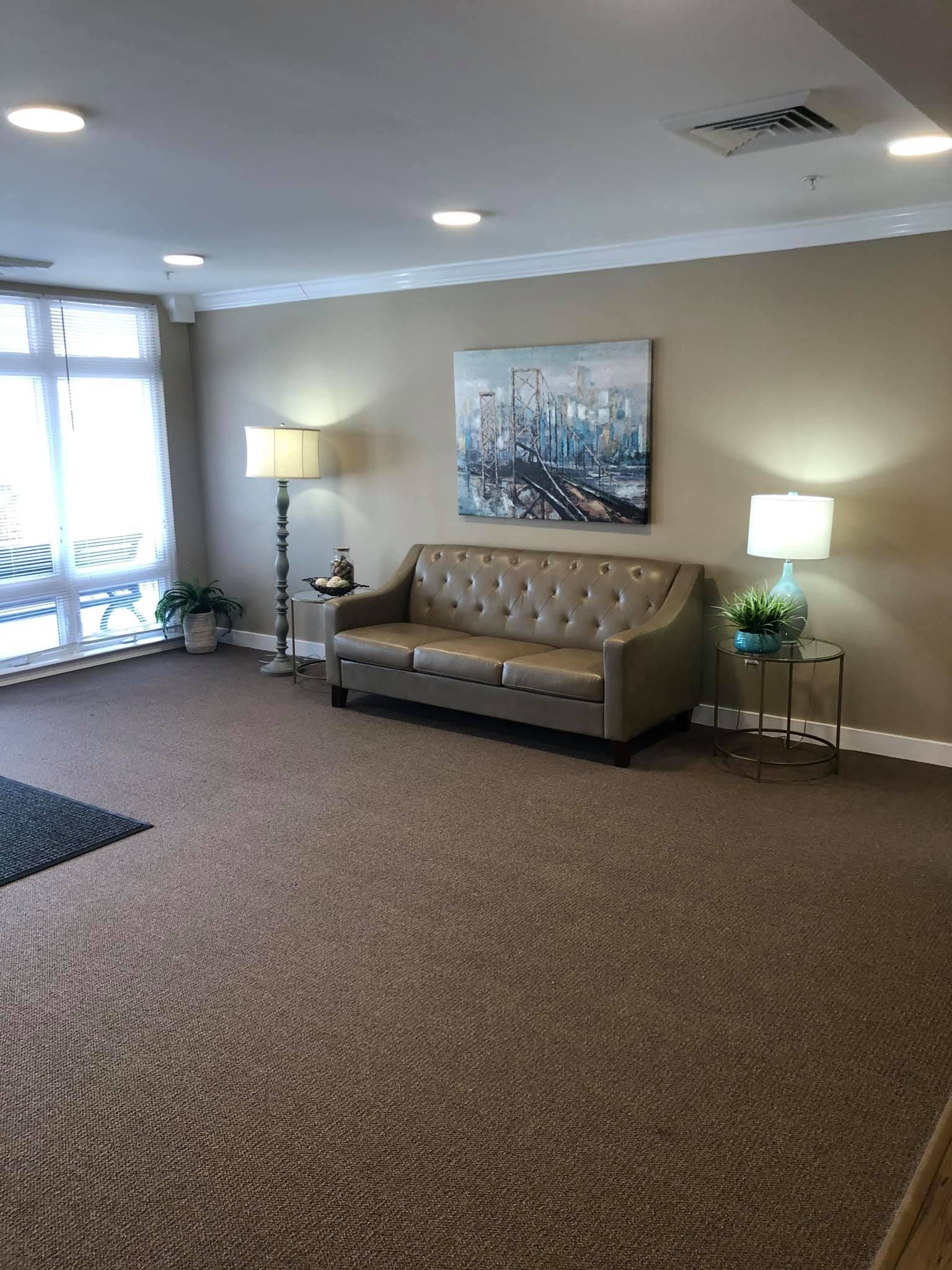A carpeted waiting area with a beige tufted sofa, a piece of city art, two lamps, and plants near a large window.
