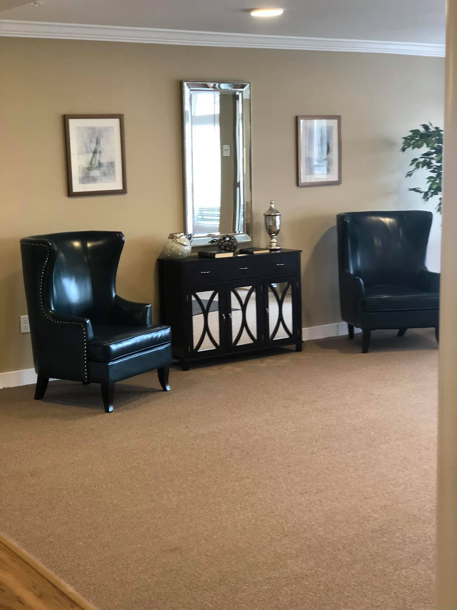 A room with two black leather wingback chairs flanking a black sideboard with a mirror and framed art on a beige wall.