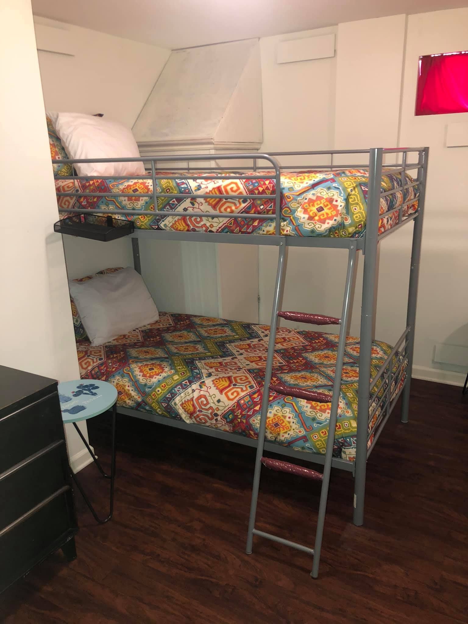A metal bunk bed with colorful patterned bedding sits in a room with white walls and dark wood floors.