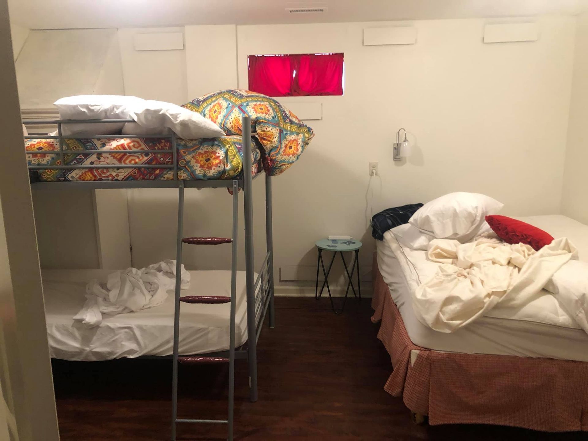 A bedroom with a metal bunk bed on the left and a queen-sized bed on the right, featuring white linens and a small table.