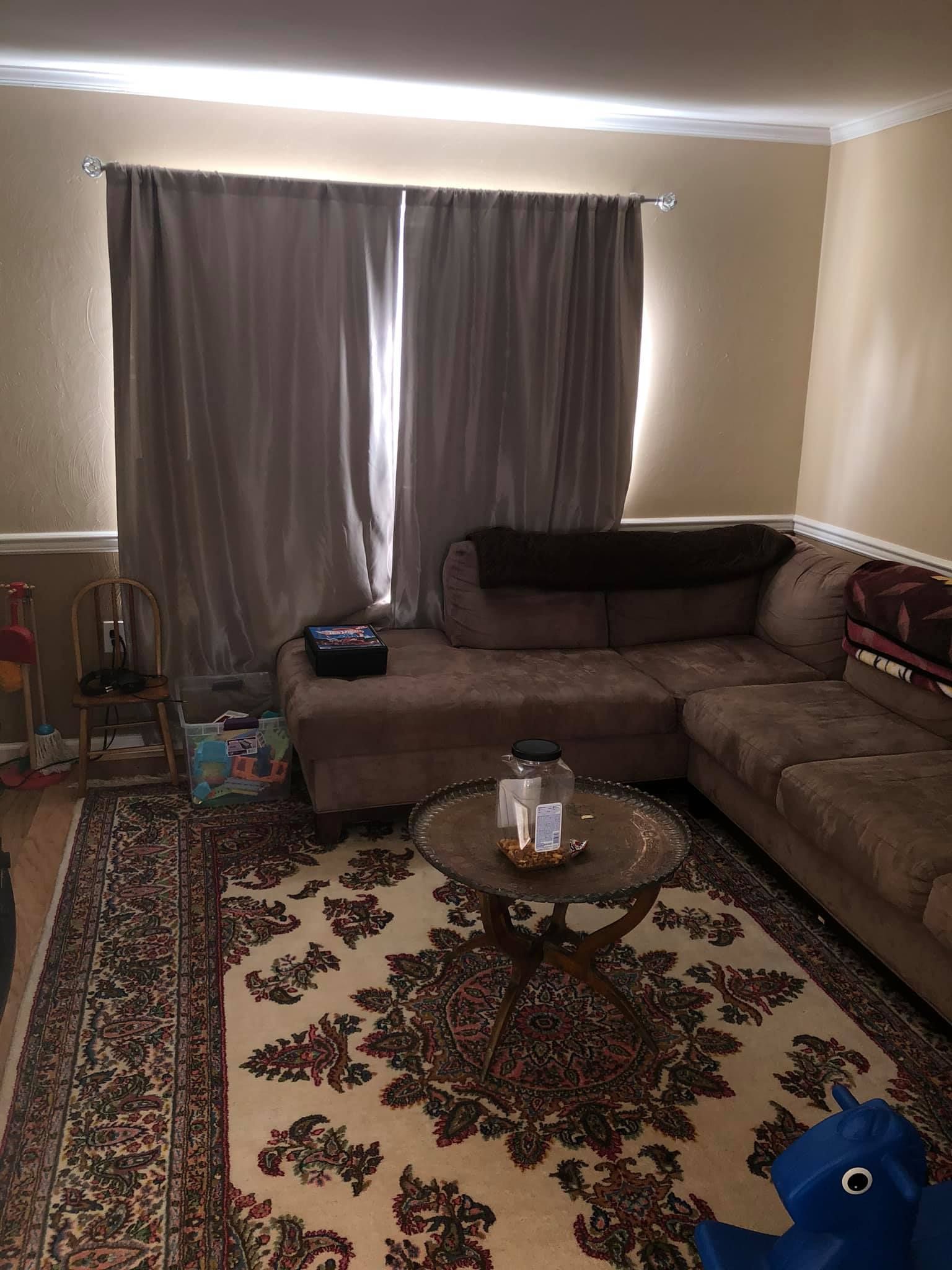 A living room featuring a brown sectional sofa, a round glass-top coffee table, and patterned rug under beige curtains.