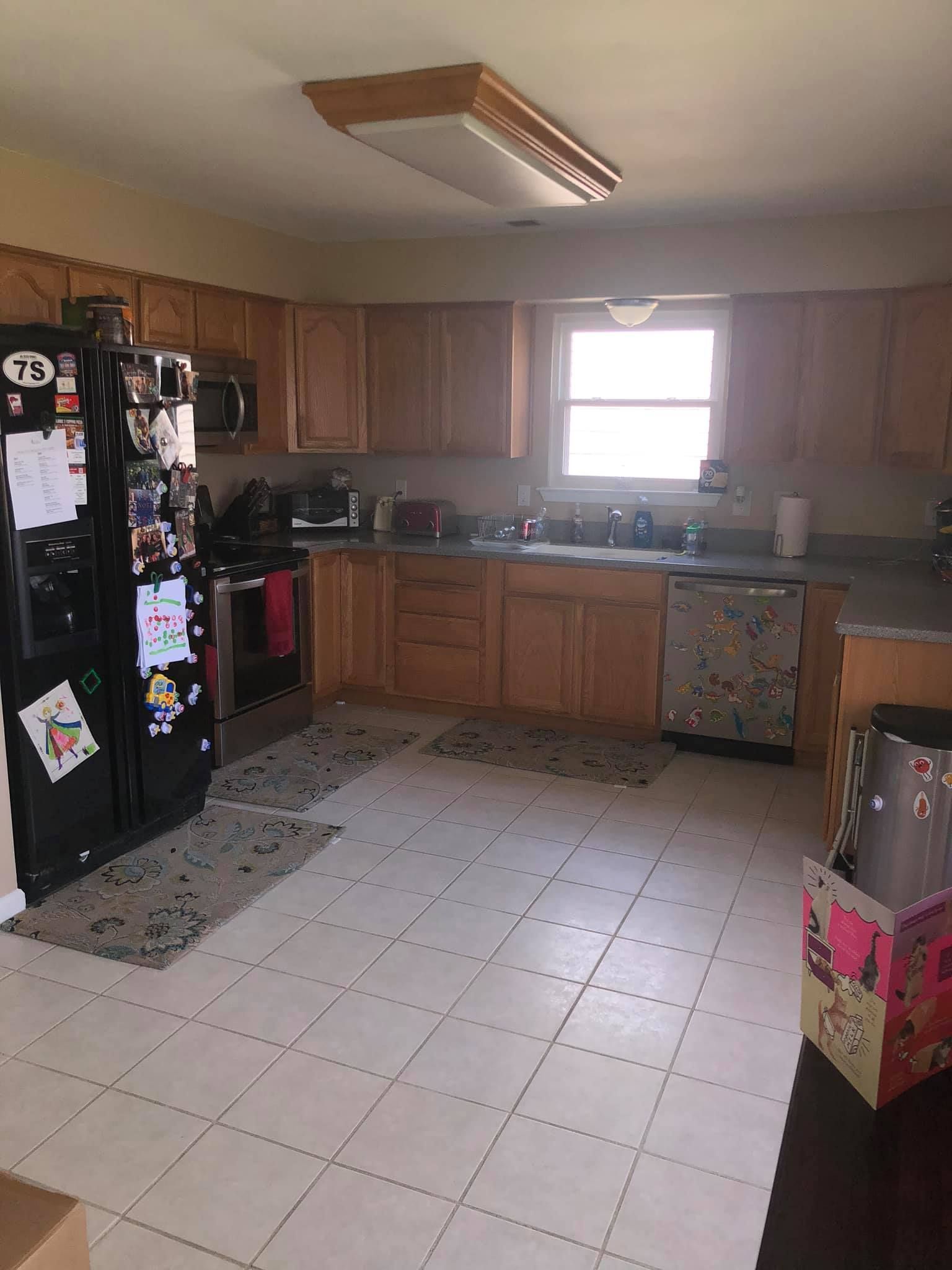 A residential kitchen featuring wooden cabinetry, a black refrigerator, white tiled floor, and a window above the sink.