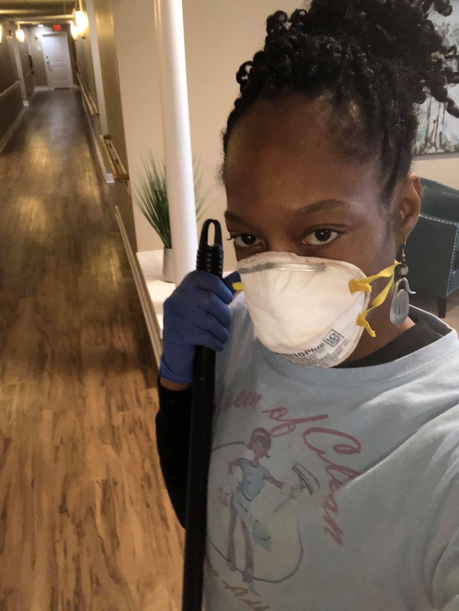 A person wearing a face mask and blue gloves holds a cleaning tool in a bright hallway.