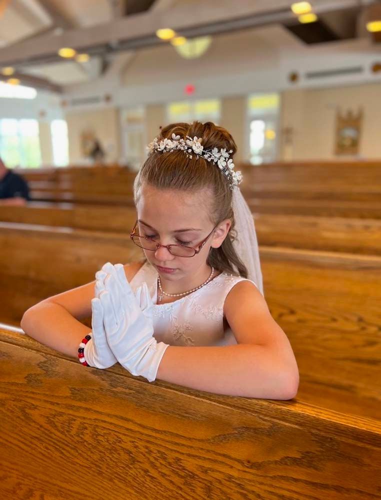 A little girl in a white dress and white gloves is praying in a church.