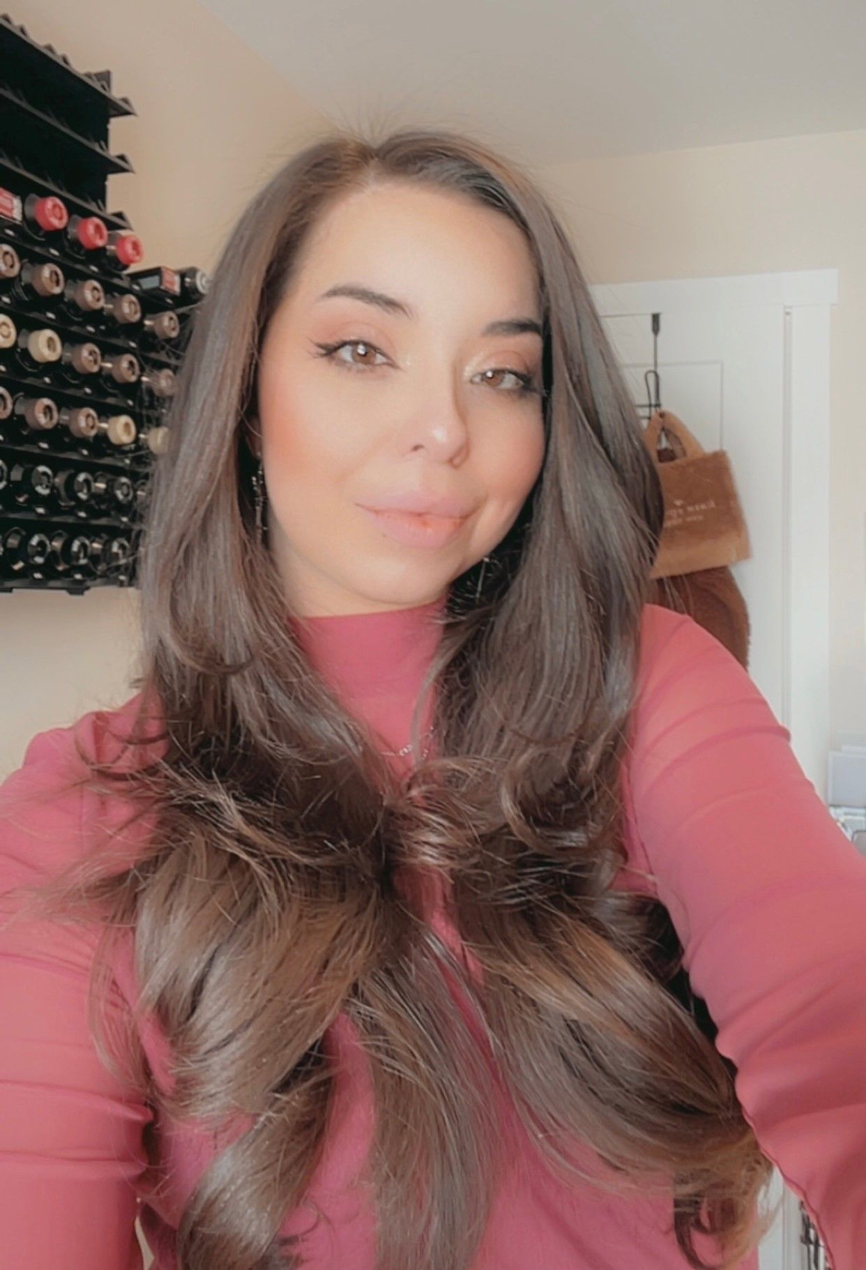 A woman with long brown hair is taking a selfie in front of a wine rack.