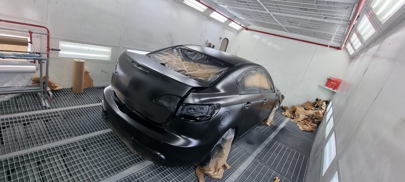 A black car inside a paint booth with parts covered.