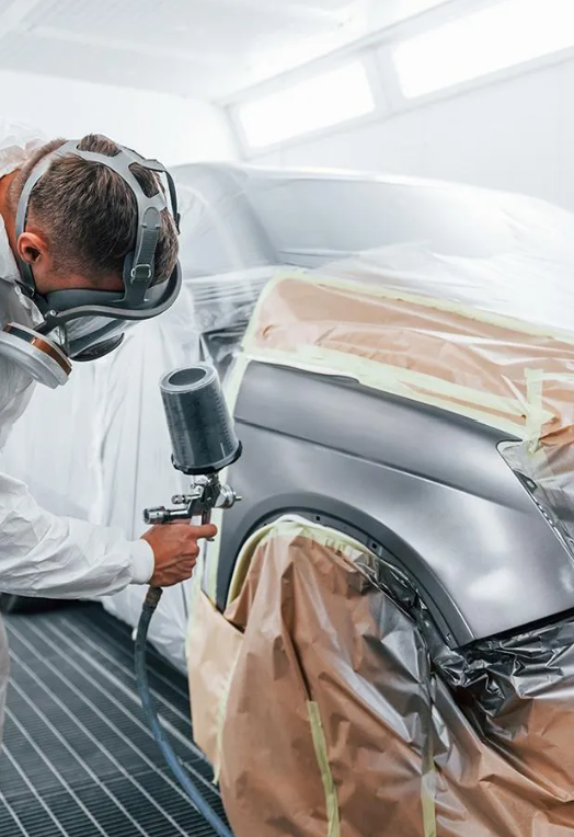 Person in protective suit using spray gun to paint a car fender.