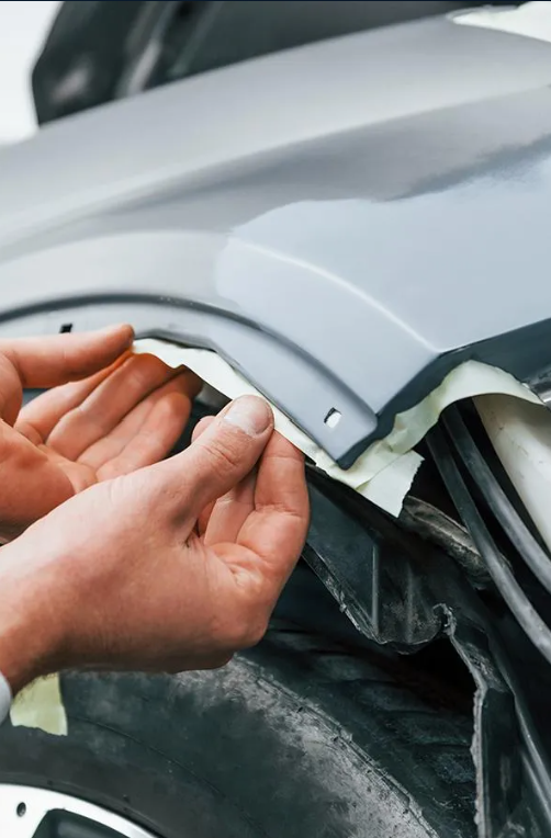 Hands applying masking tape to a car's painted body panel in a repair shop.