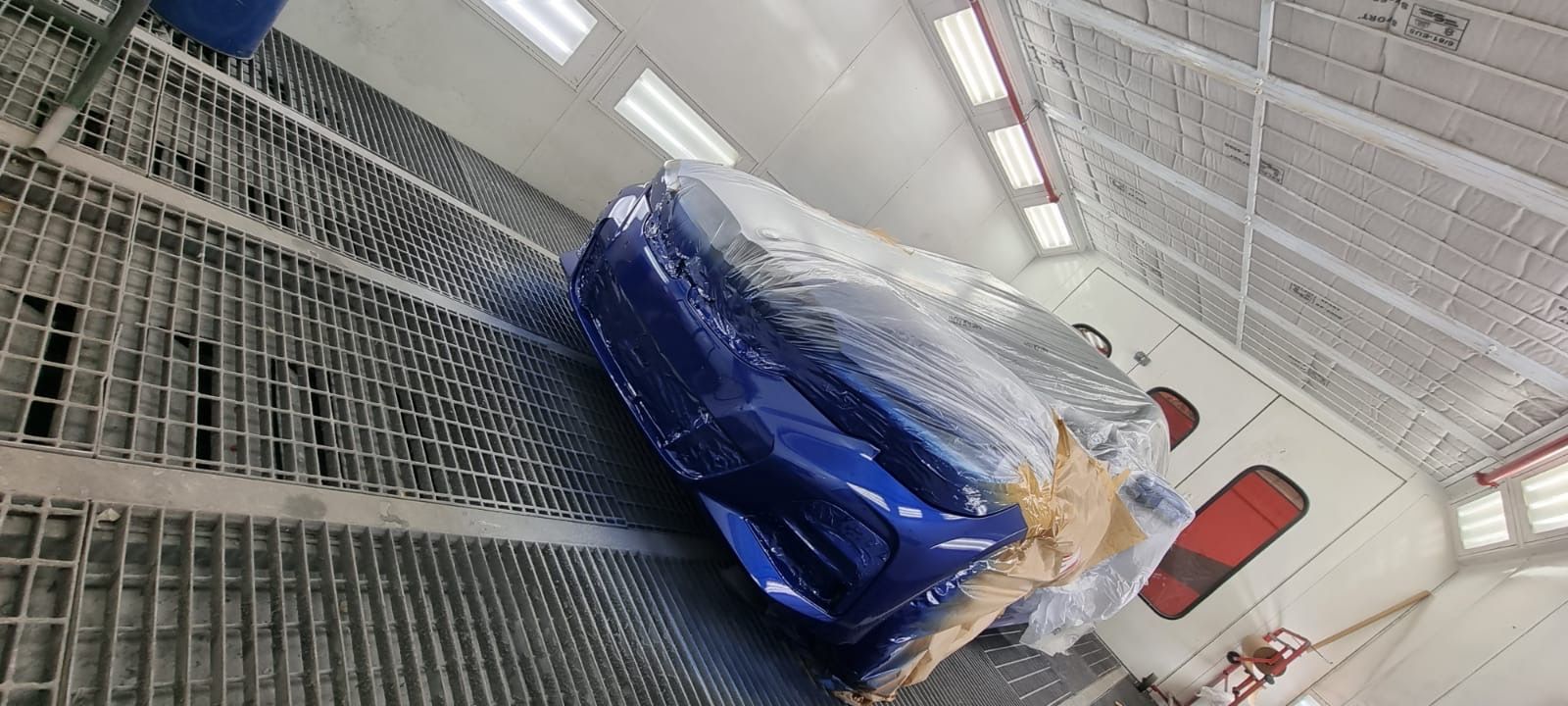 Blue car in a paint booth covered in plastic wrap, sitting on a metal grating floor.