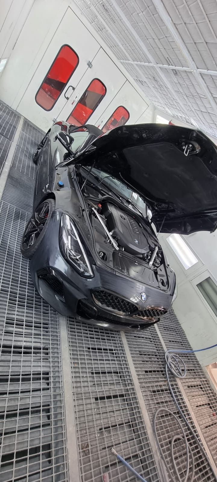 A dark gray sports car with its hood open inside a paint booth with open overhead lighting.