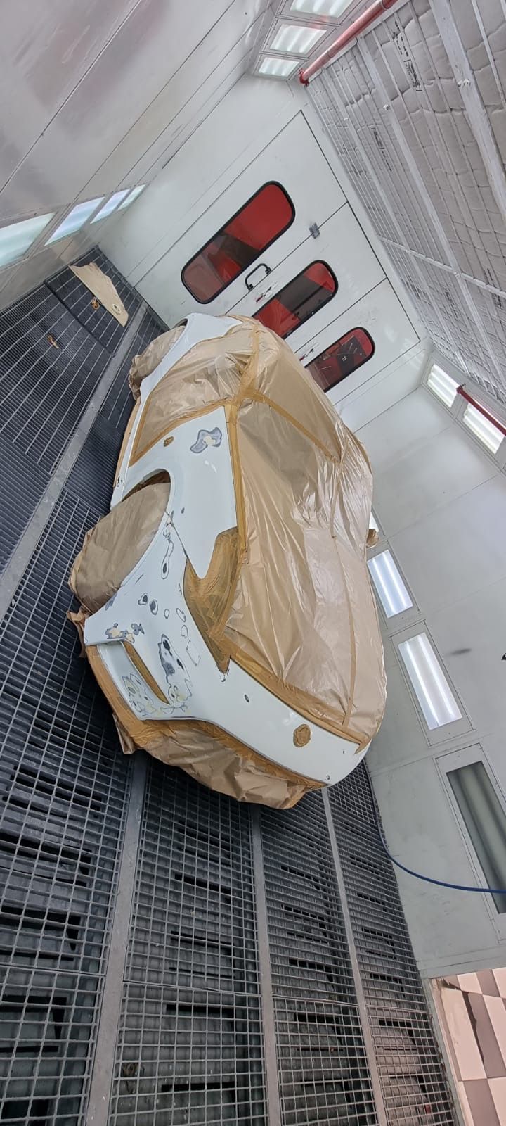 A car part wrapped in brown paper inside a spray paint booth with a metal grate floor and white walls.