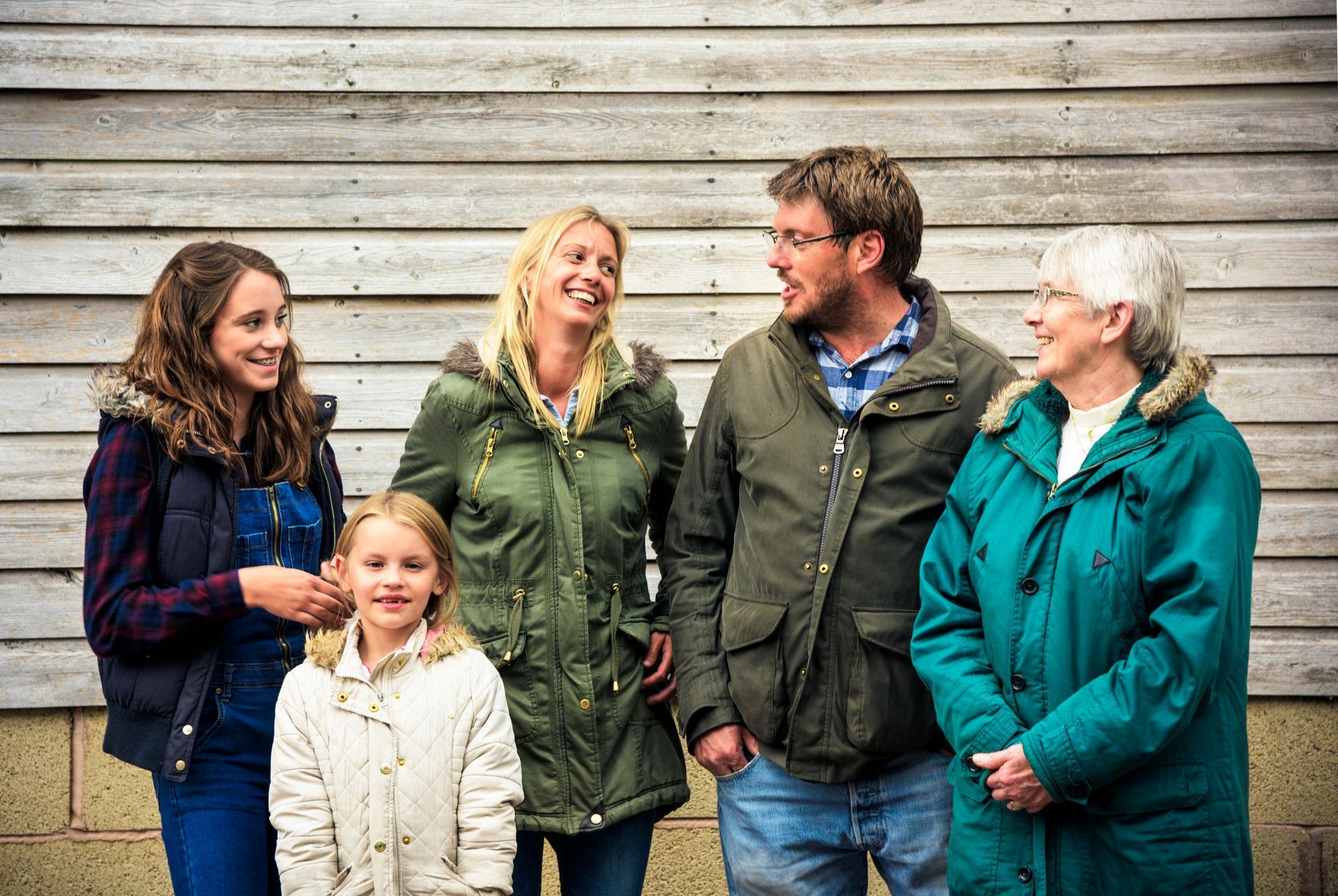 A family of five stands in front of a wooden wall, smiling and laughing. They are dressed in outdoor attire.