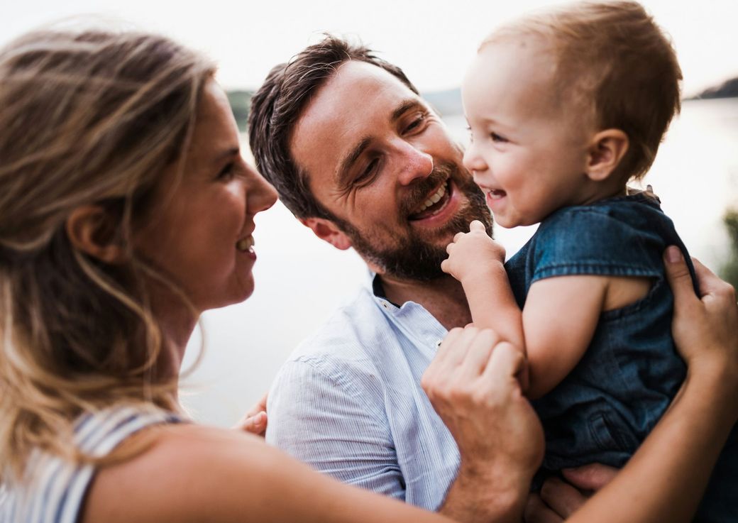 Parents smile at a toddler, who is being held. The family is outside near water.