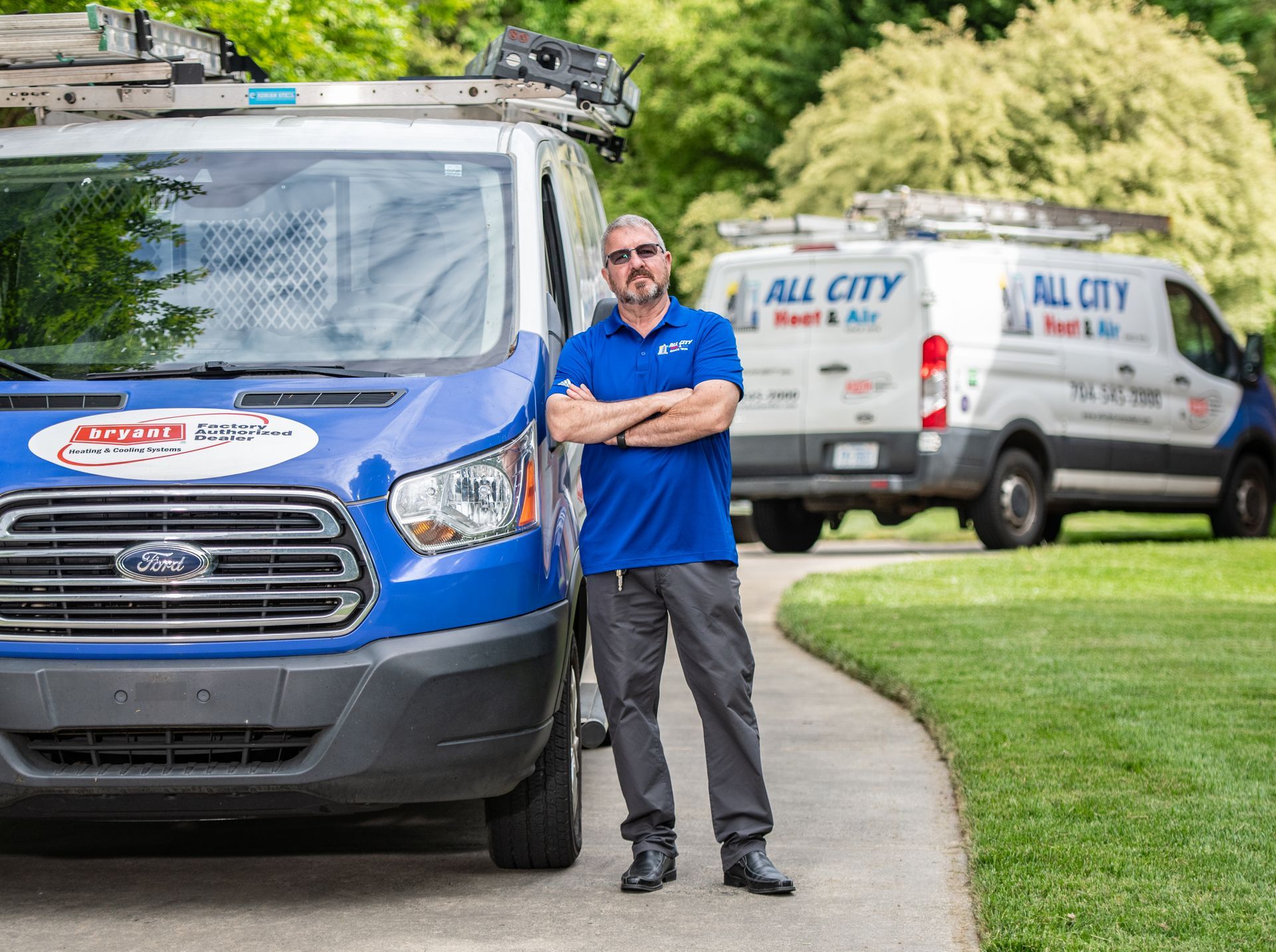 An All City Heat and Air technician is standing in front of two All City Heat and Air vans.