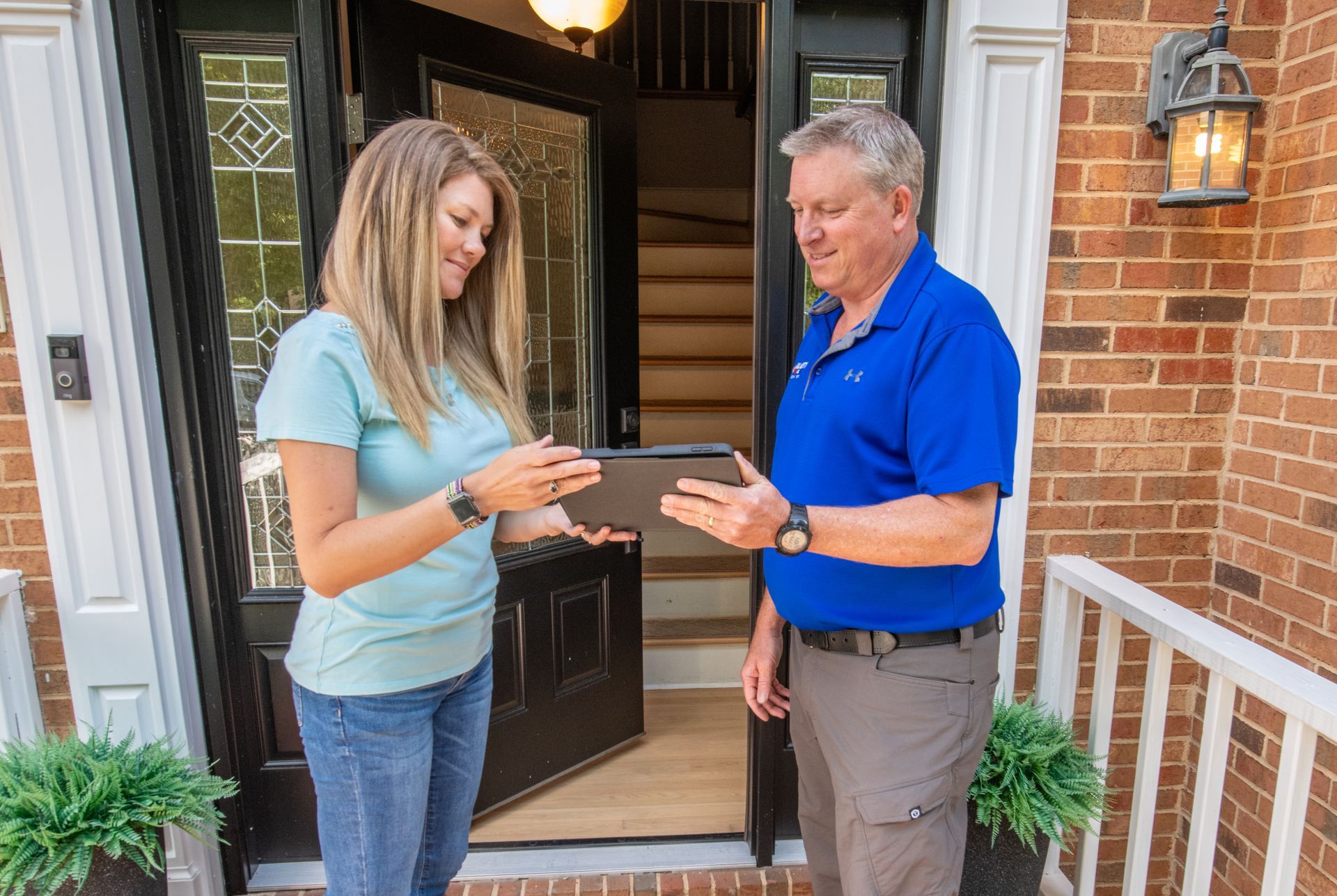 An All City Heat and Air technician and a woman are standing in front of a brick house looking at a tablet.