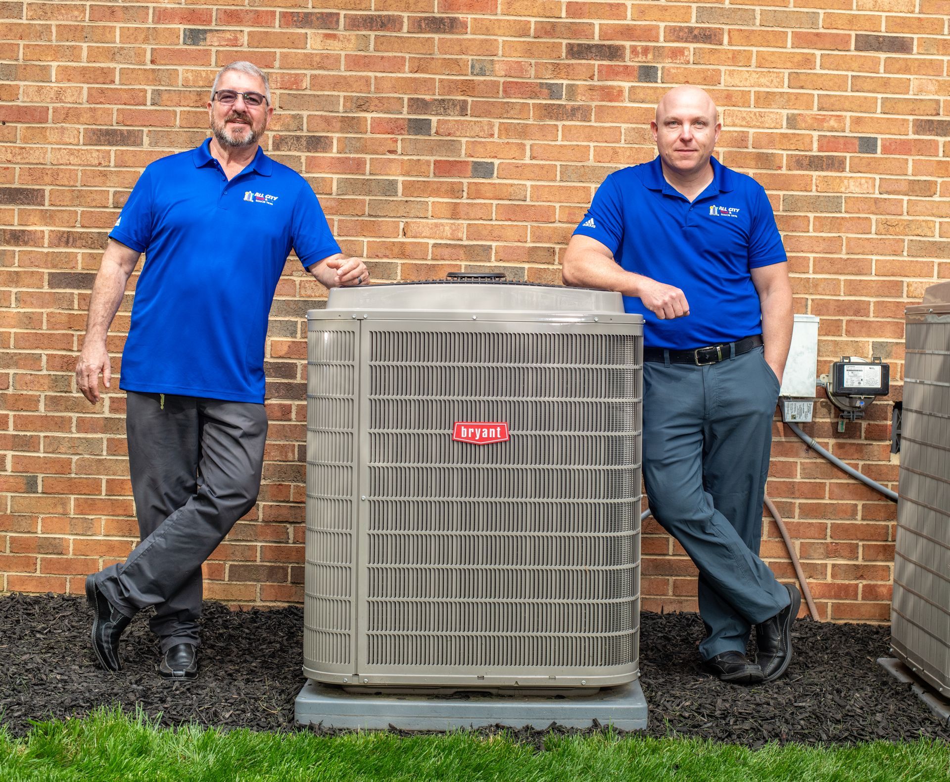Two All City Heat and Air technicians in blue shirts are standing next to an air conditioner.