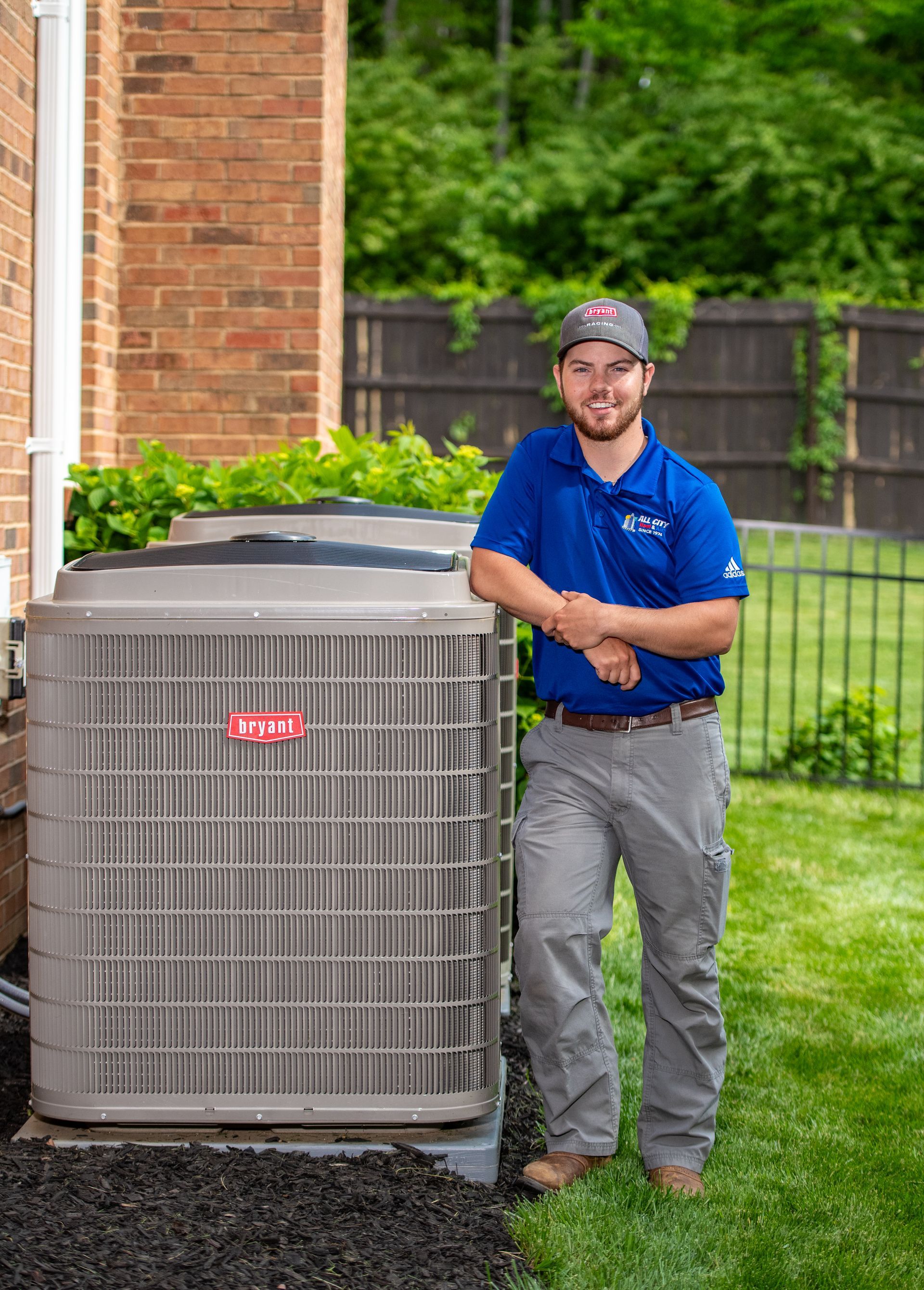 An All City Heat and Air technician in a blue shirt is standing next to an air conditioner.