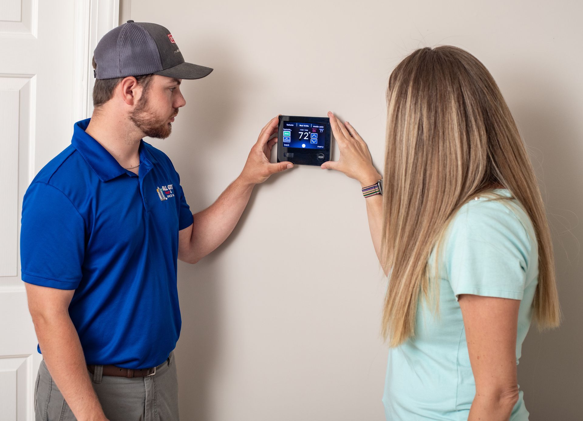 An All City Heat and Air technician and a woman are looking at a thermostat on a wall.