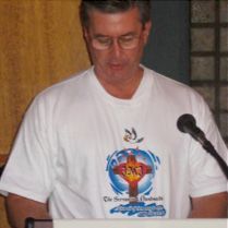A man in a white t-shirt is standing at a podium with a microphone.