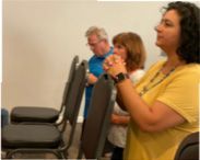 A woman in a yellow shirt is praying in front of a group of people sitting in chairs.