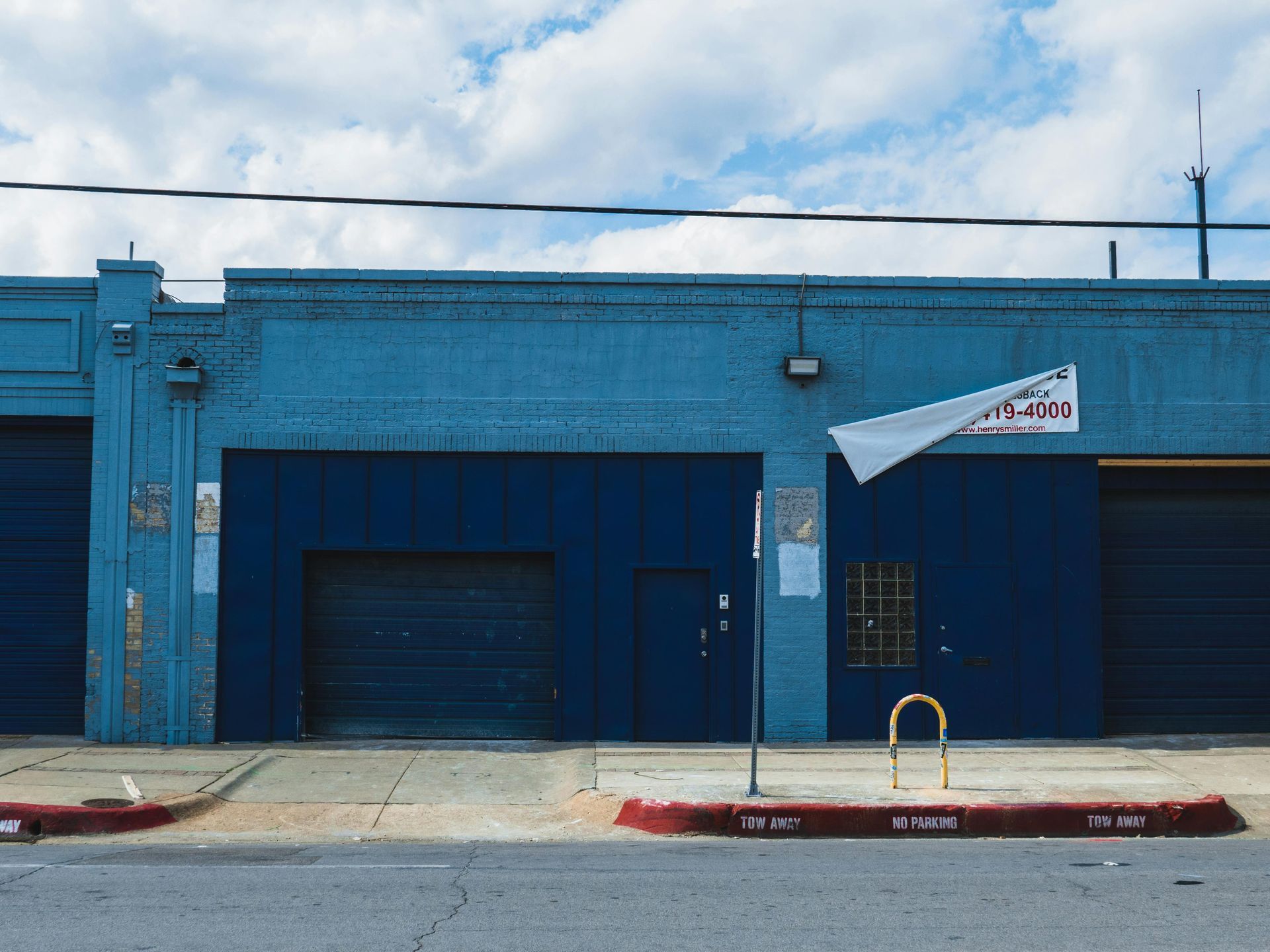 Blue building with garage doors, street and sidewalk. A sign hangs from the building.