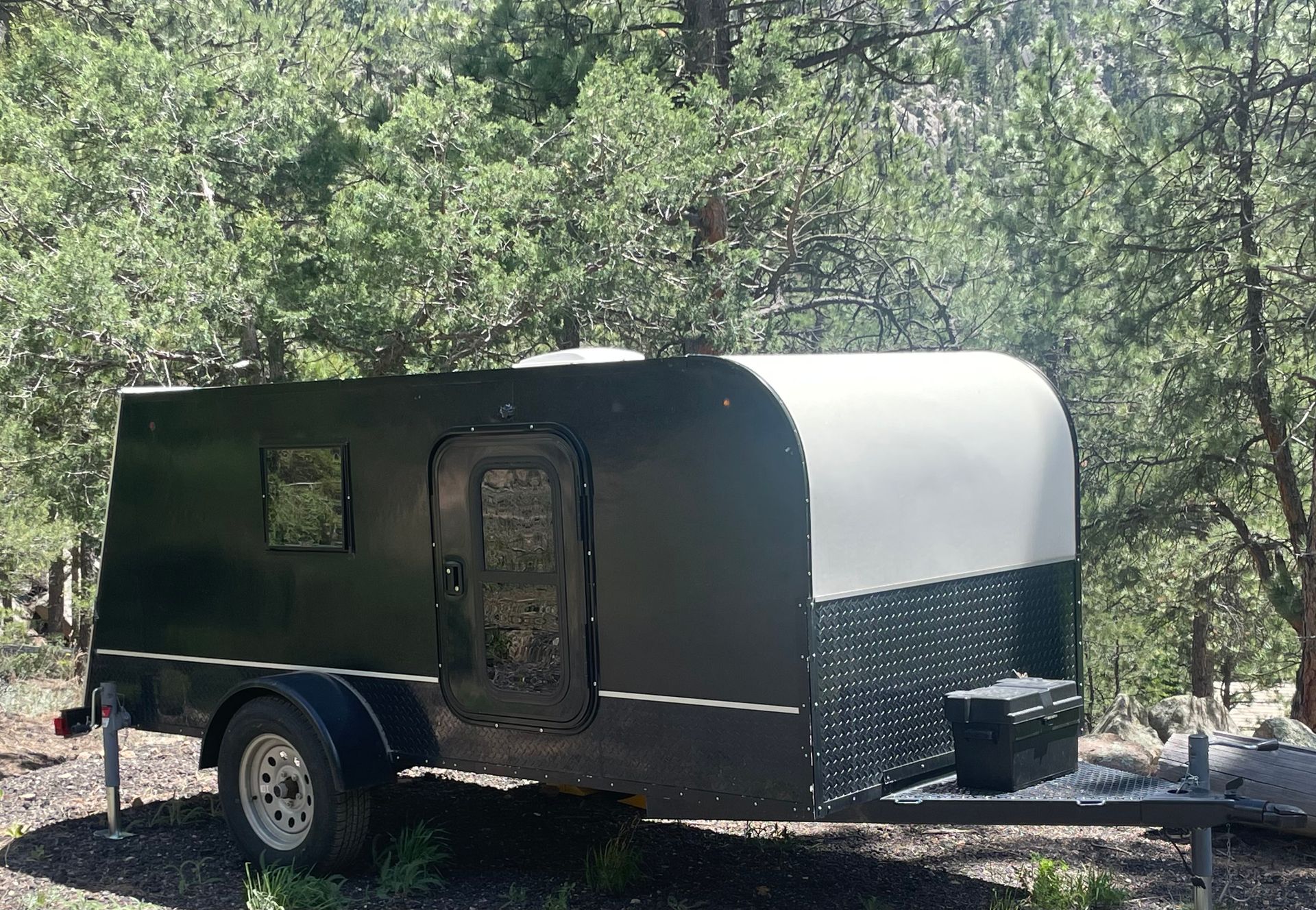 Black and gray camper trailer parked outdoors in front of trees.