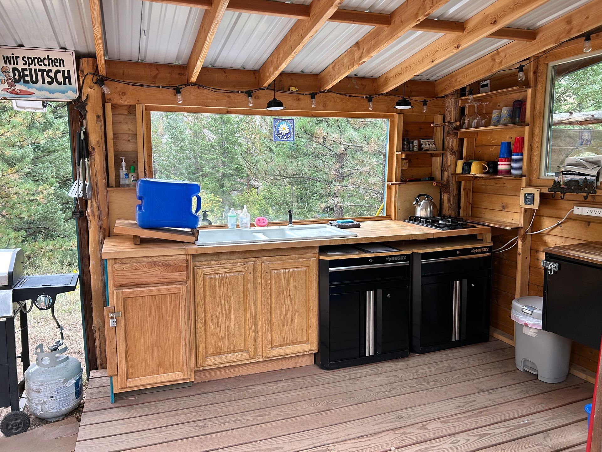 Outdoor kitchen with wooden cabinets, appliances, and a large window overlooking trees.