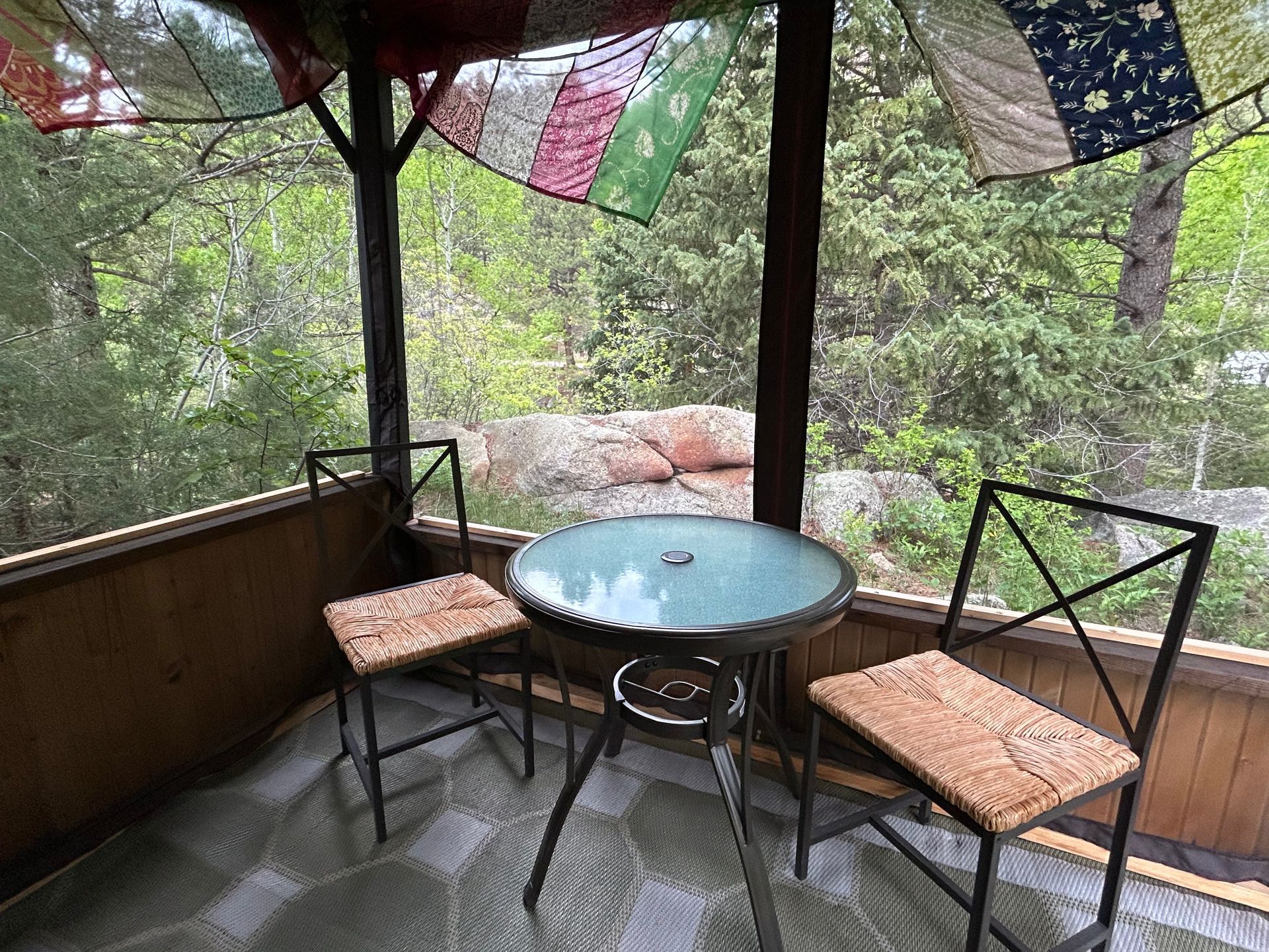 Screened-in porch with a glass-topped table and two chairs, overlooking a forest. Colorful prayer flags hang above.