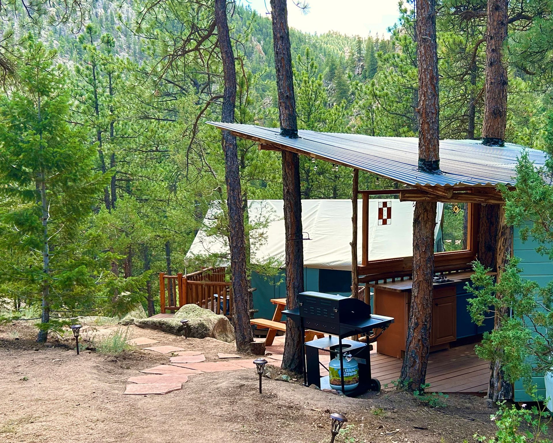 Outdoor cooking area with grill, picnic table, and tent in a wooded area.