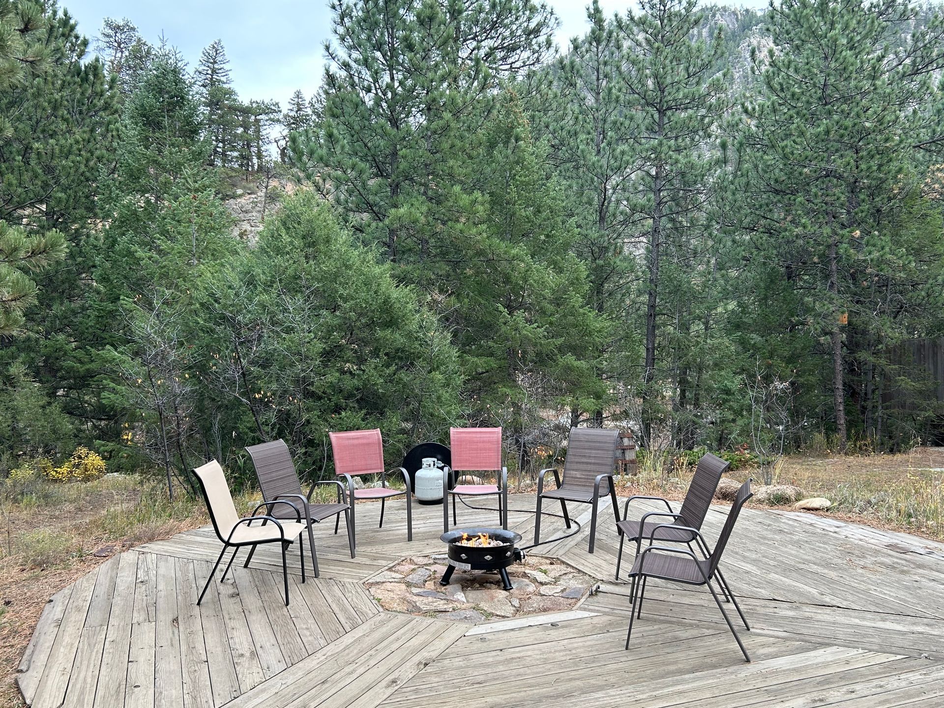 A circle of chairs around a fire pit on a wooden deck in a wooded area.