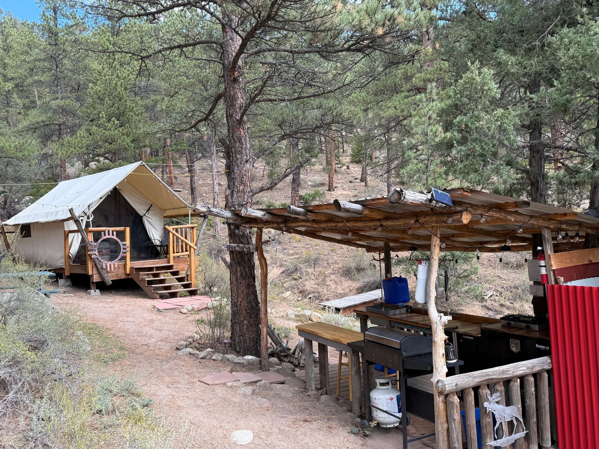 Canvas tent with a wooden deck and a rustic kitchen shelter in a wooded area.