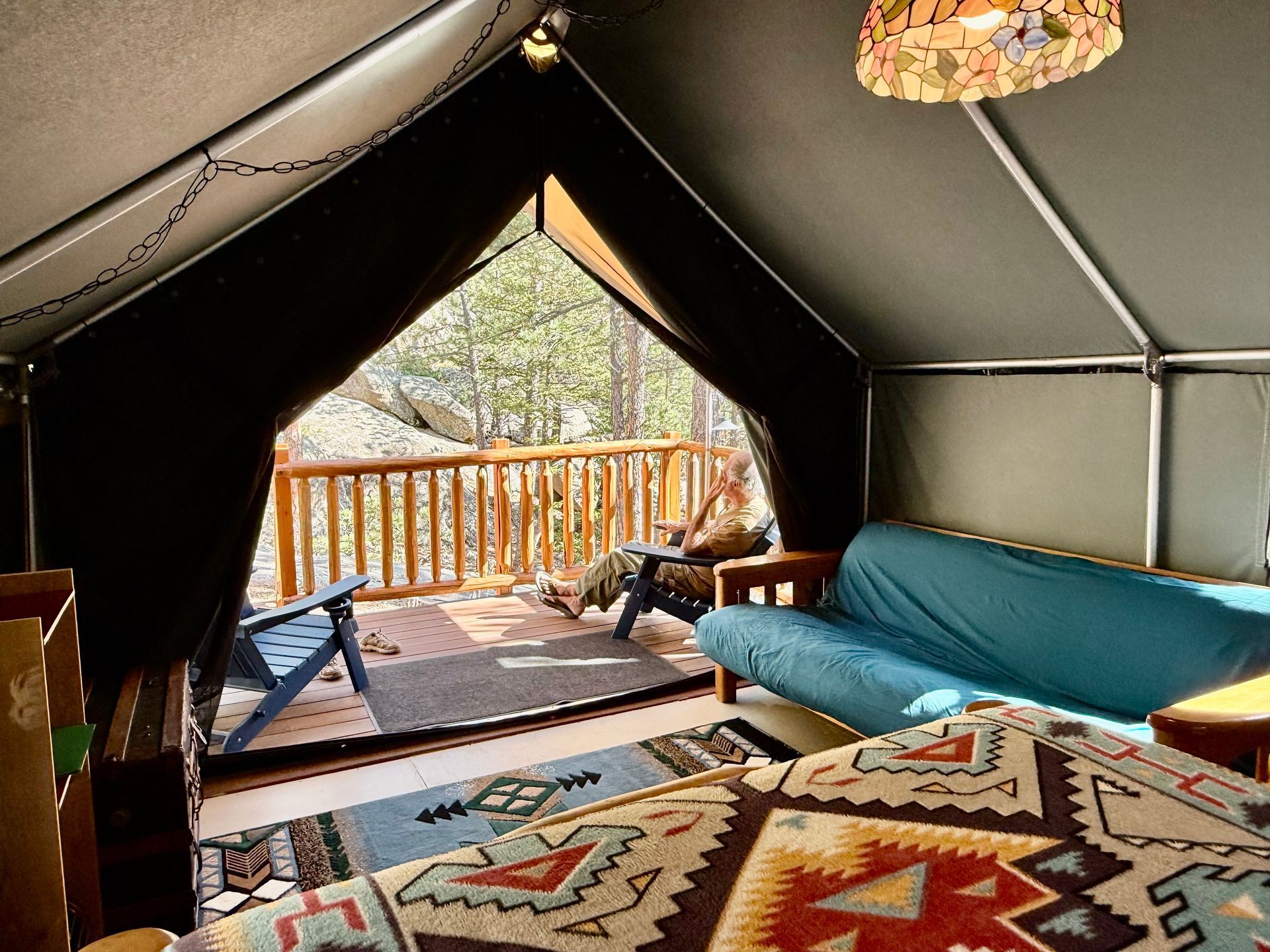 Inside of a glamping tent, open to a deck. A person sits on the deck. Colorful rugs and a blue futon inside.