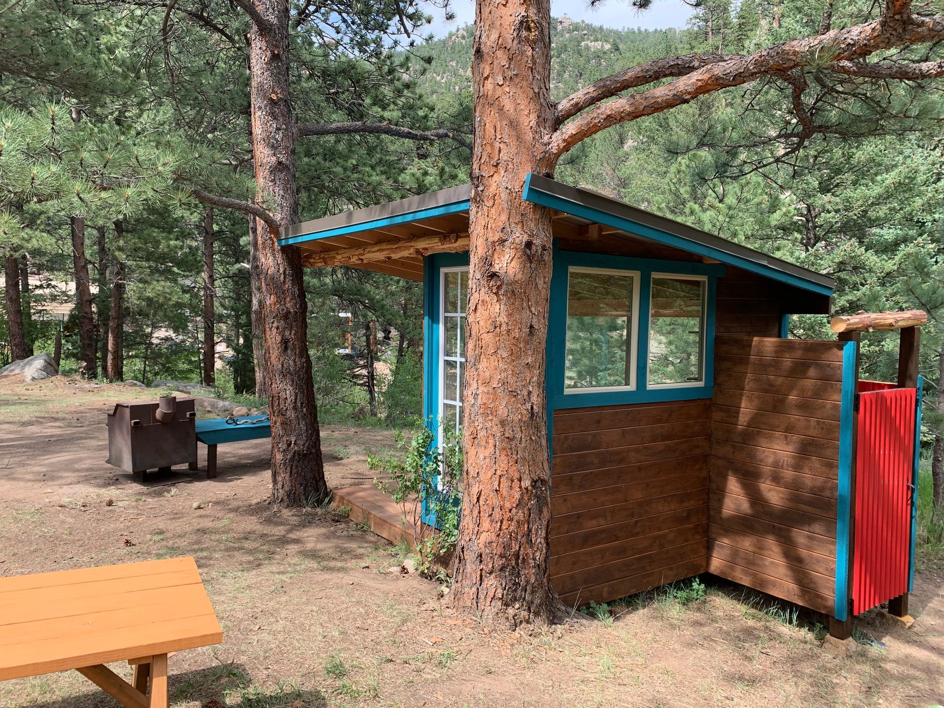 Small cabin with teal trim nestled between trees in a wooded area; outdoor seating and a table are nearby.