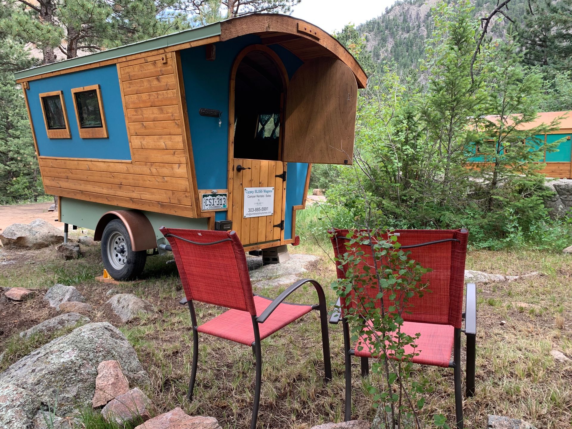 A small wooden camper on a trailer with two red chairs in front, nestled in a wooded area.