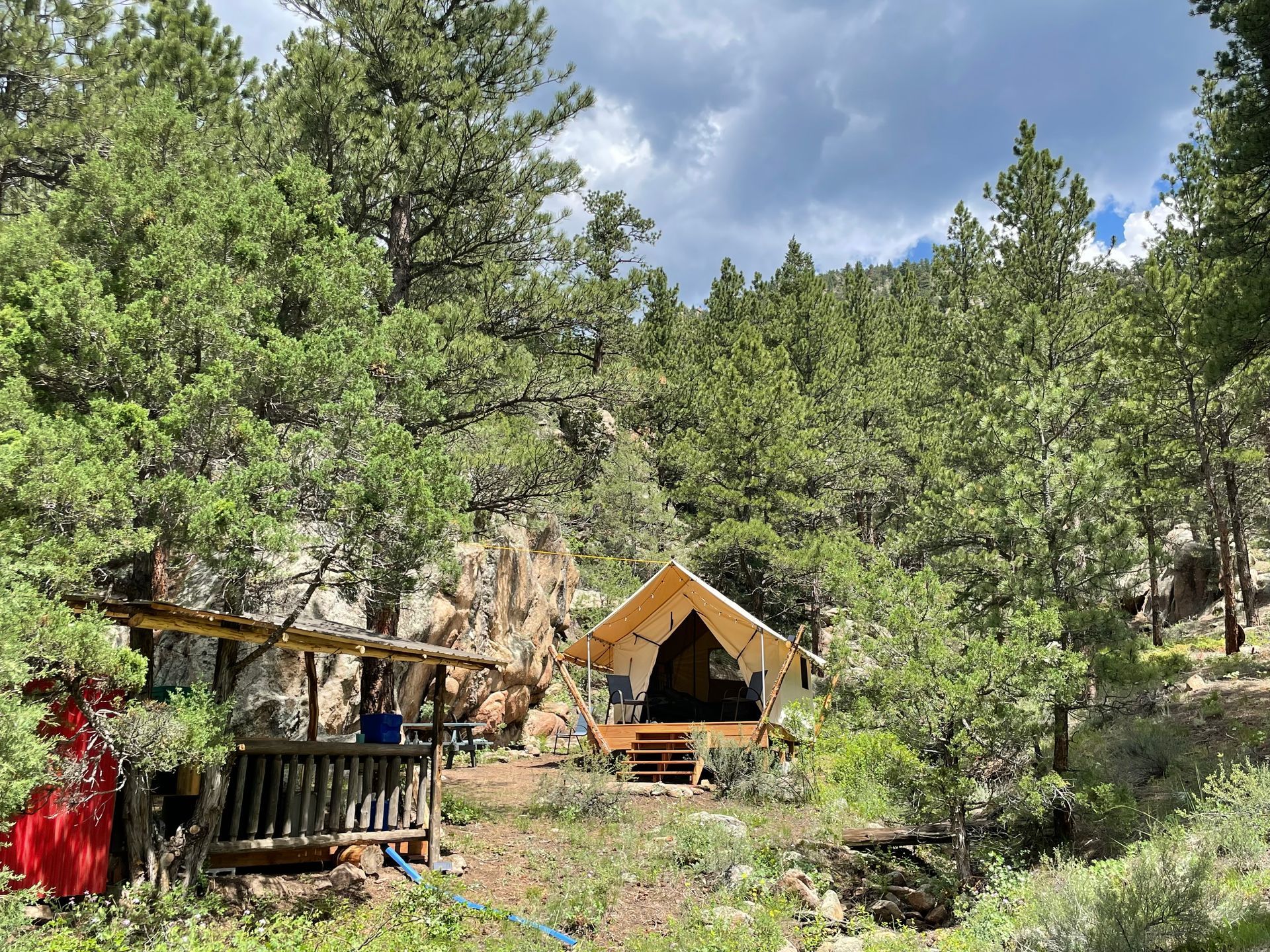 Canvas tent and small wooden deck nestled in a lush, green forest with cloudy sky above.