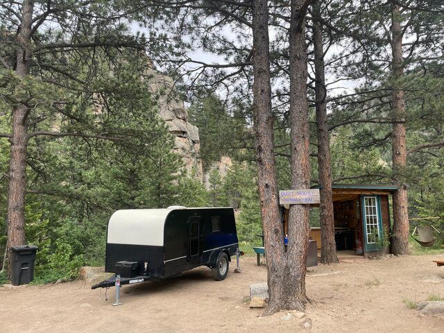 Small camper trailer parked near a small shed and trees in a forest.