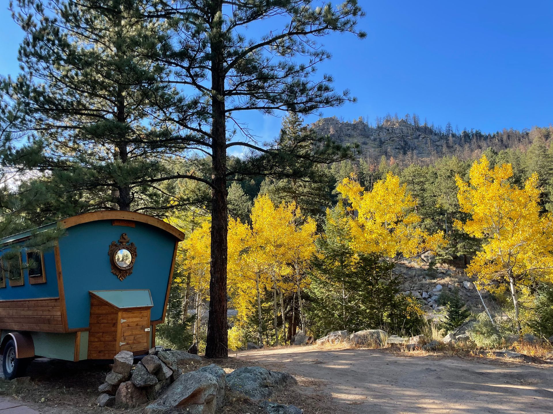 Blue camper with rounded roof in a forest setting; yellow autumn leaves, mountains in the background.
