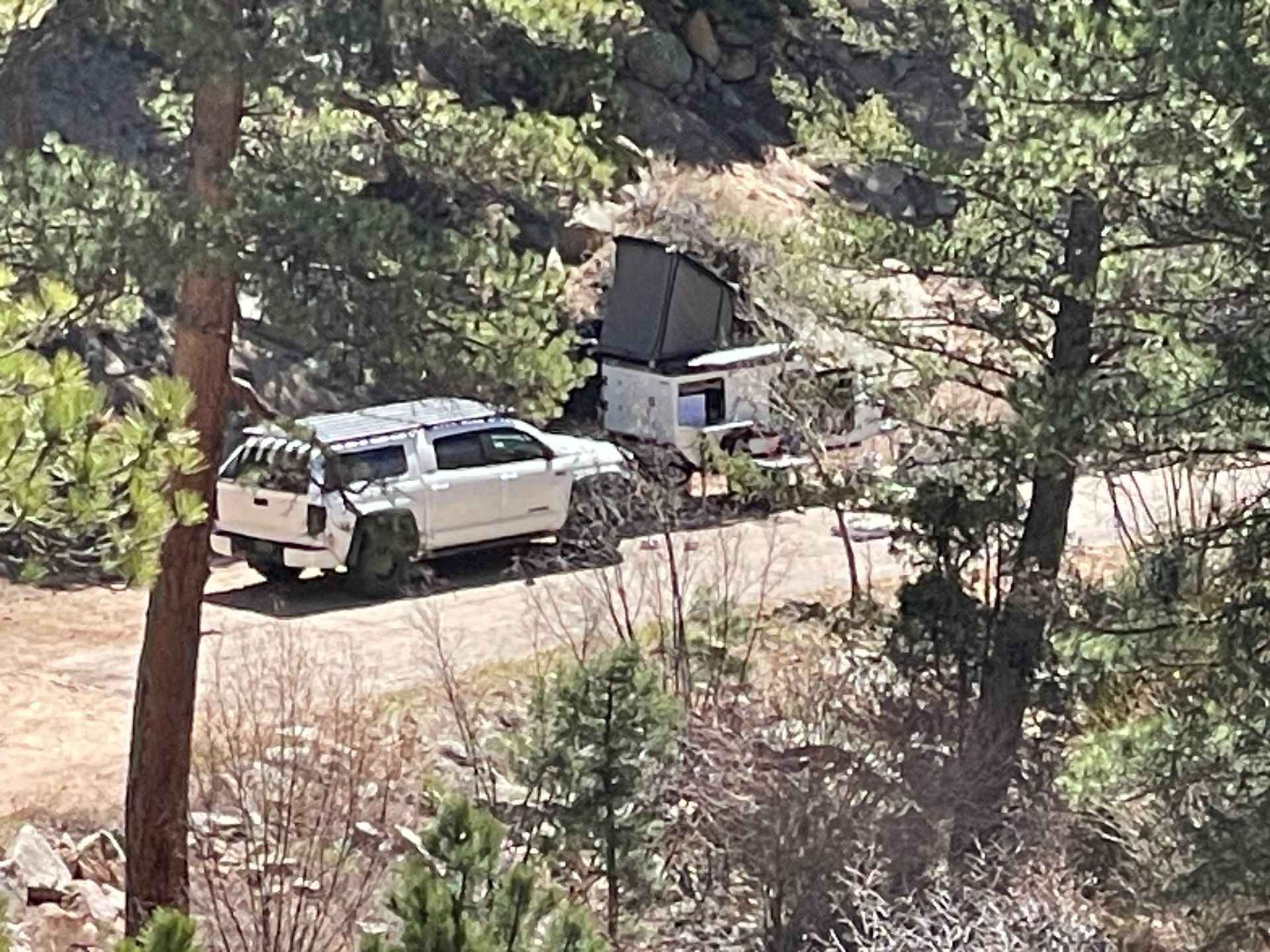 White truck and camper trailer parked on a dirt road in a wooded area.