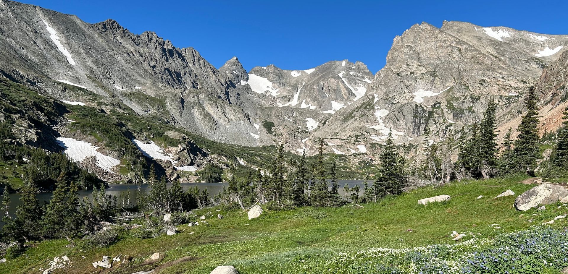 Mountain landscape with snow, trees, and a lake under a clear blue sky.